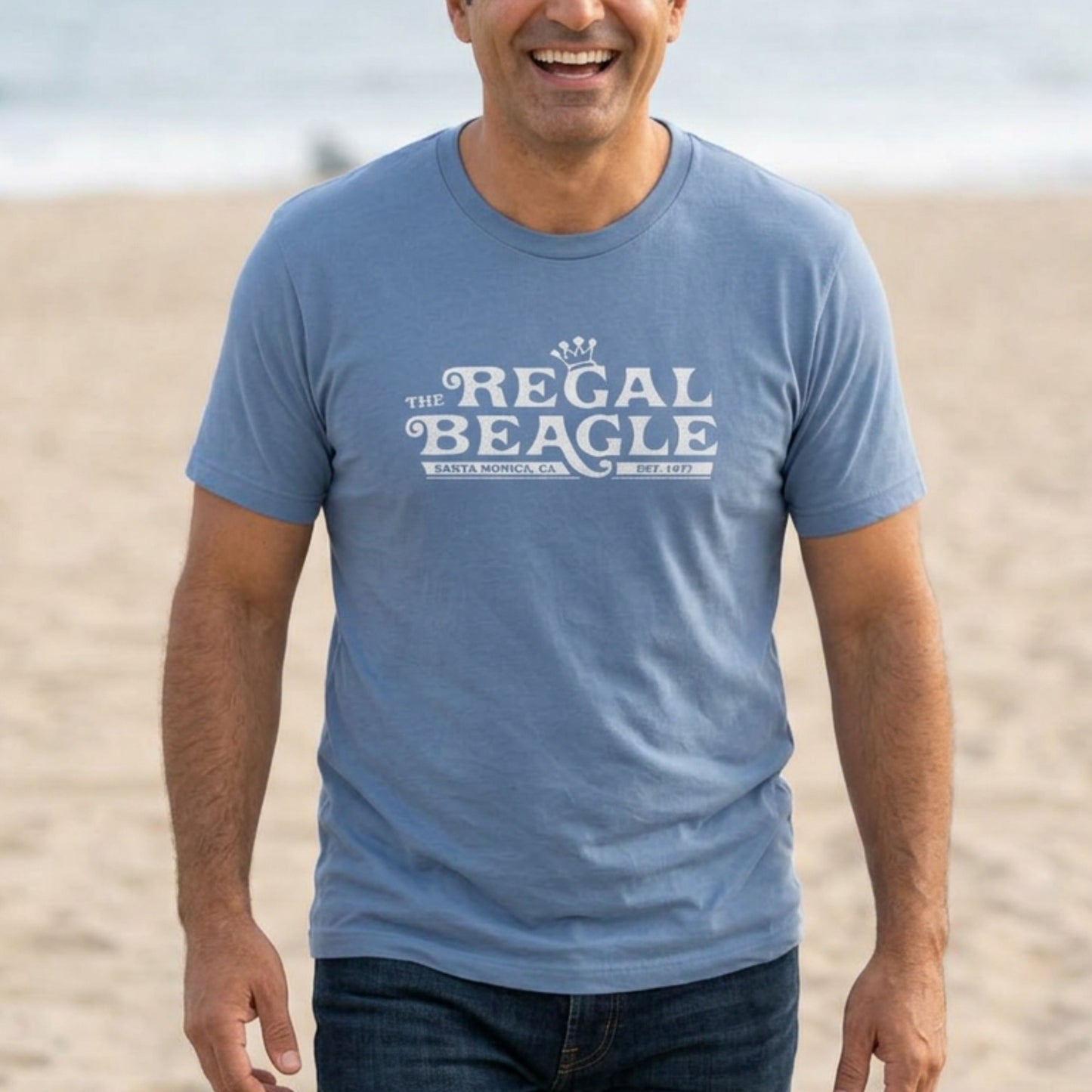 Person wearing Lavender Blue Bella+Canvas t-shirt with the Regal Beagle logo from the TV Show 'Threes Company' in a vintage distressed style.