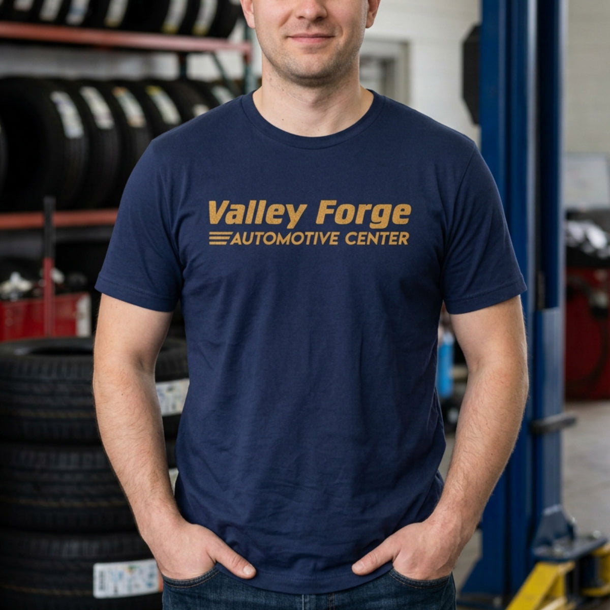 Person in an auto shop wearing Navy tee with vintage gold Valley Forge Automotive Center auto shop logo from comedian Shane Gillis Tires TV show