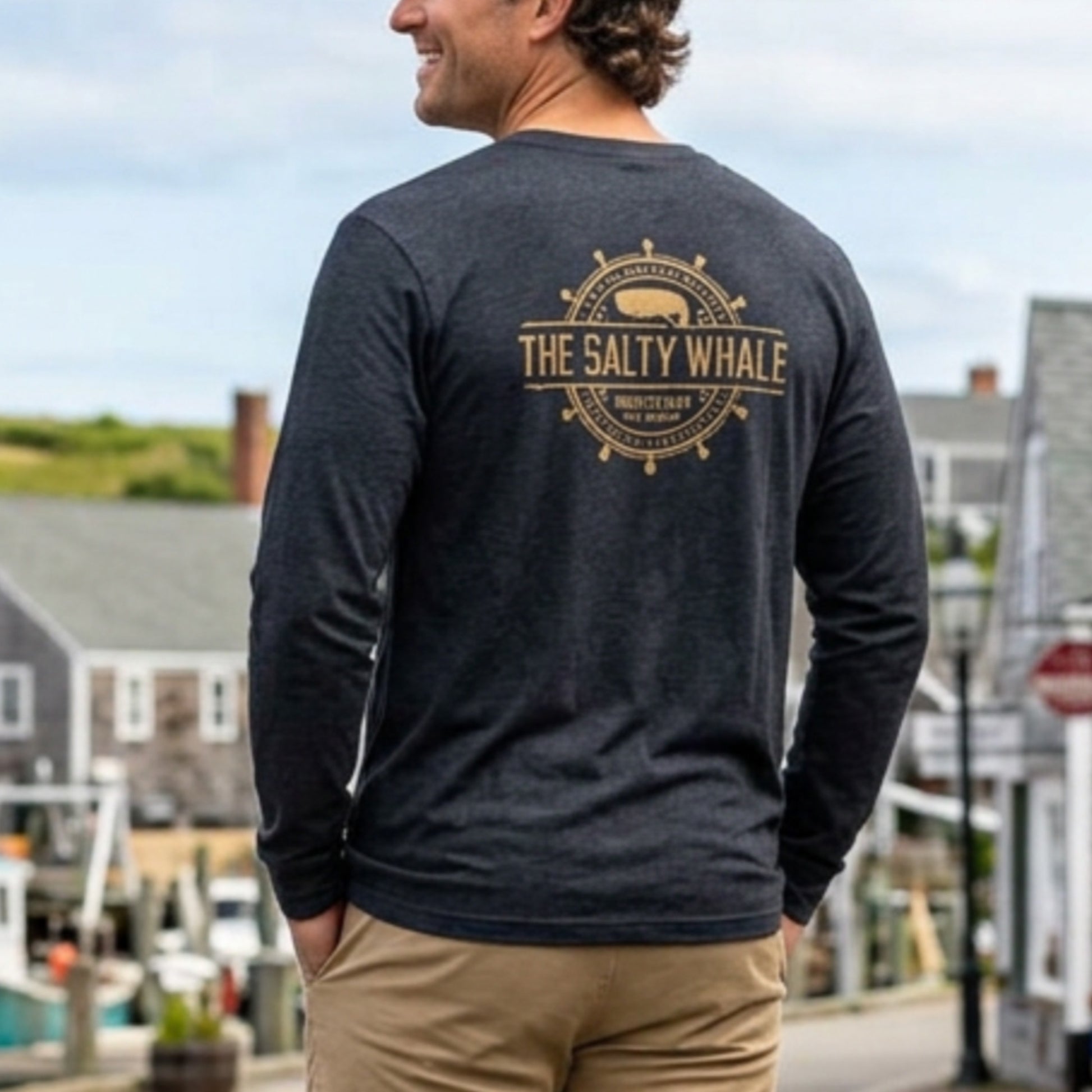 Back of person wearing Heather navy blue long sleeve tee with vintage gold The Salty Whale logos from the TV show Widow's Bay nautical on front and back