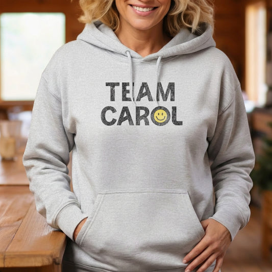 Woman wearing Ash unisex Heavy Blend pullover hoodie sweatshirt with TEAM CAROL vintage distressed smiley graphic
