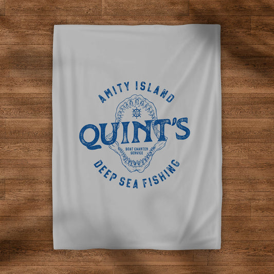 Gray velveteen plush blanket with vintage blue Quint’s Deep Sea Fishing logo inspired by Jaws, shown on a wood floor.