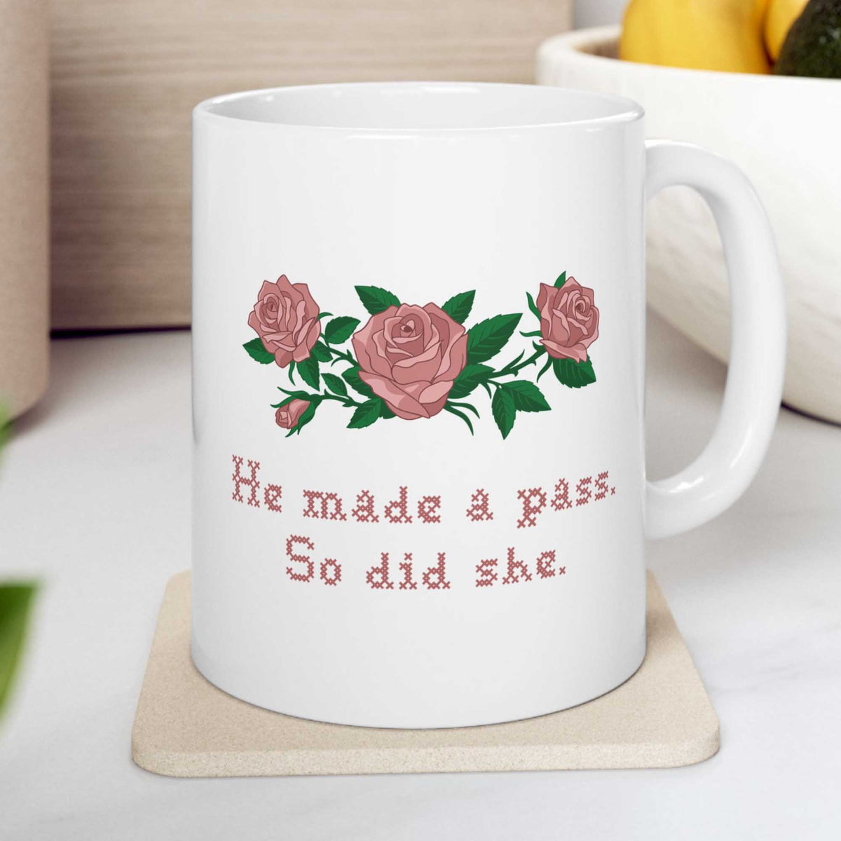 White mug on a coaster with pink roses and He made a pass. So did she. in cross stitch, inspired by Ruth in Widow's Bay