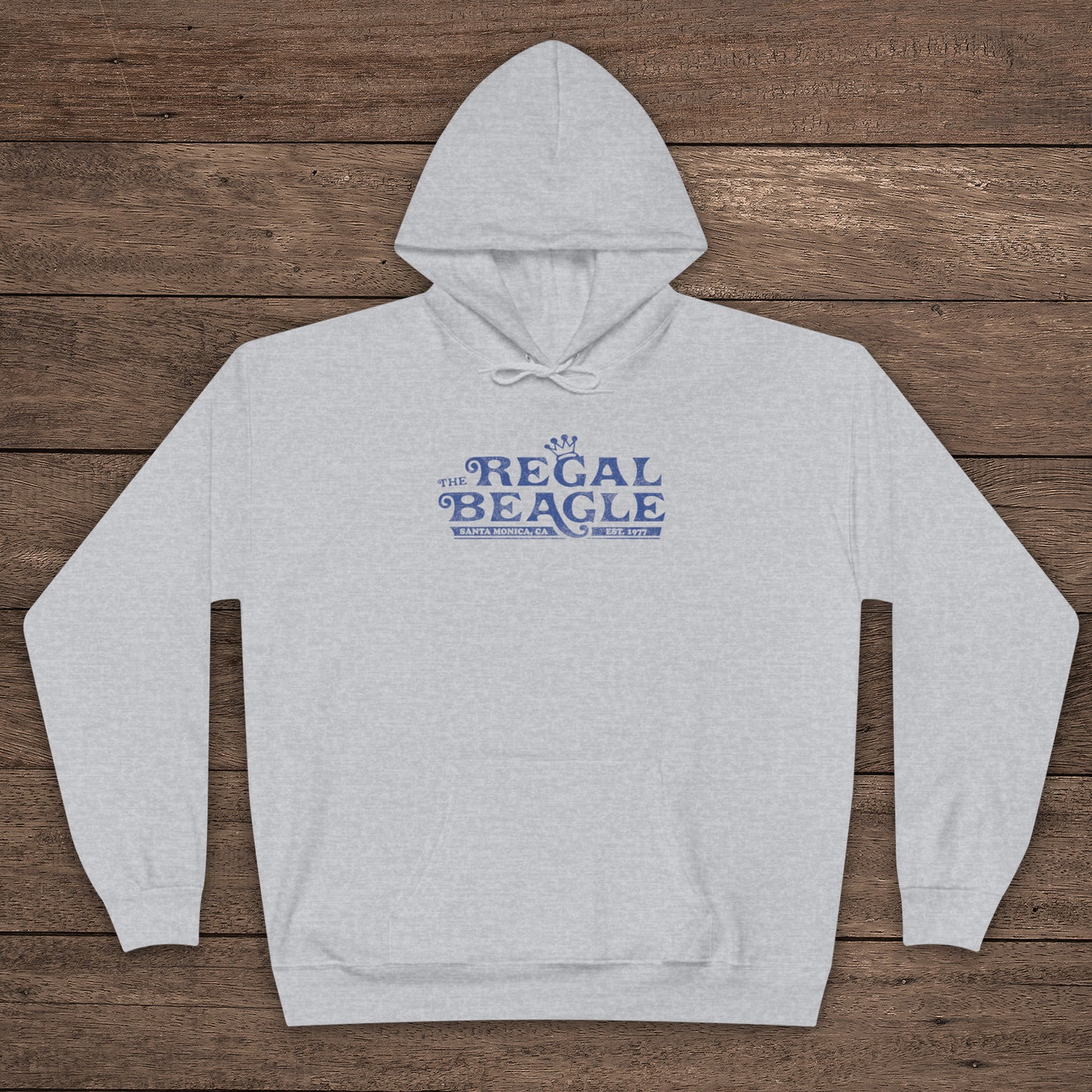 Ash gray Hanes EcoSmart pullover hoodie with vintage distressed Regal Beagle logo from Three’s Company on chest, shown on wood floor.
