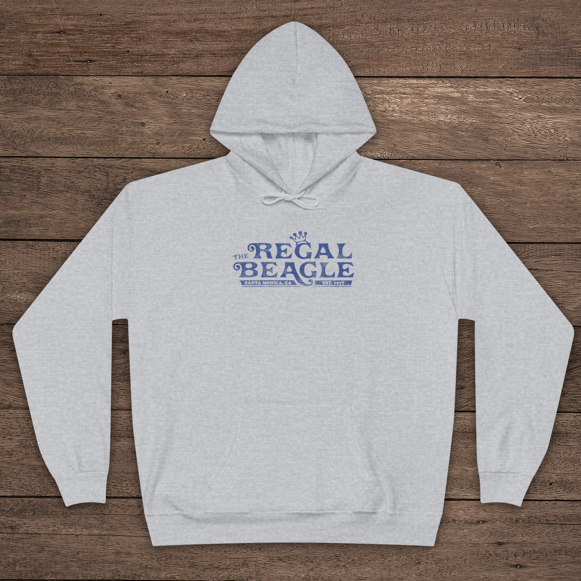 Ash gray Hanes EcoSmart pullover hoodie with vintage distressed Regal Beagle logo from Three’s Company on chest, shown on wood floor.