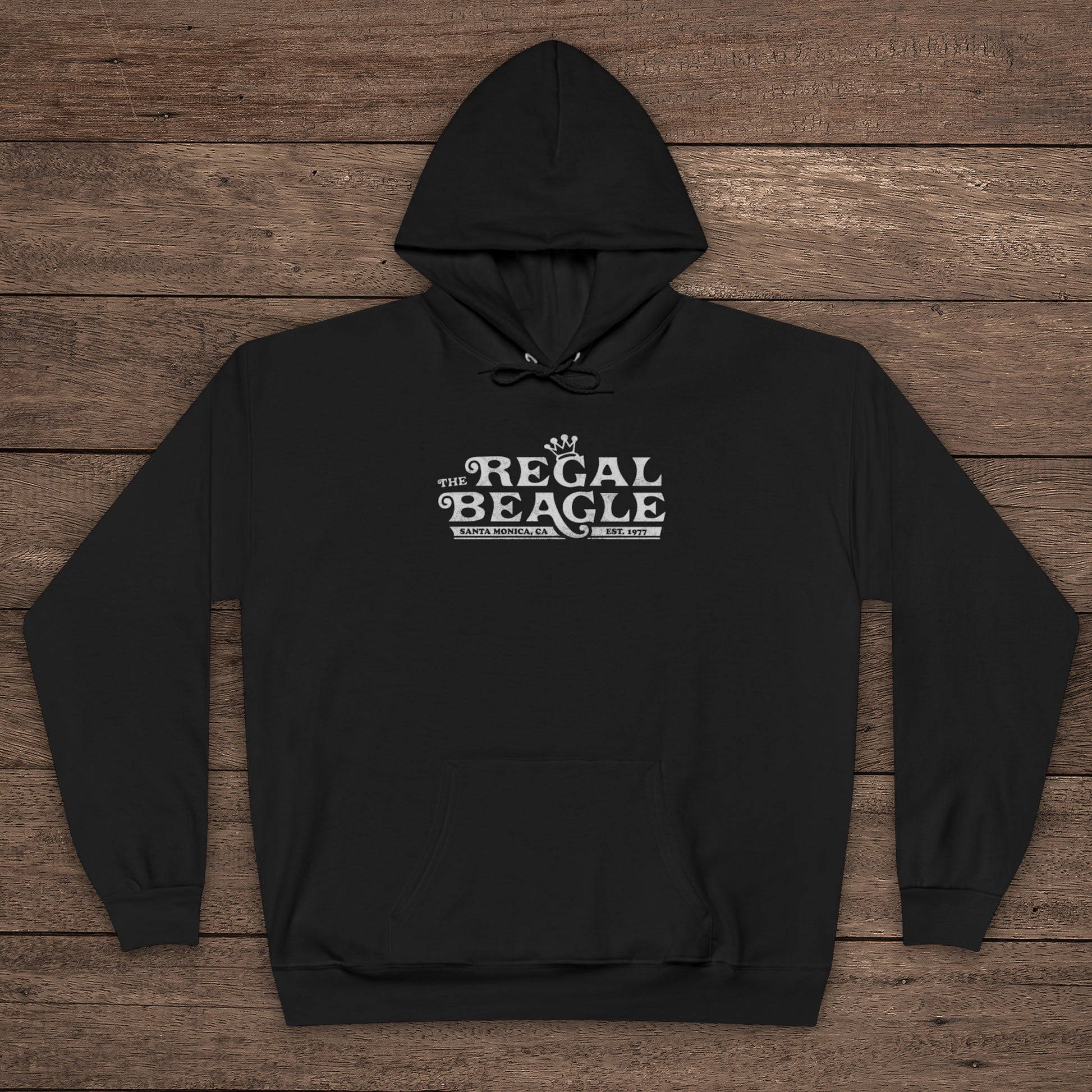 Black Hanes EcoSmart pullover hoodie with vintage distressed Regal Beagle logo from Three’s Company on chest, shown on wood floor.