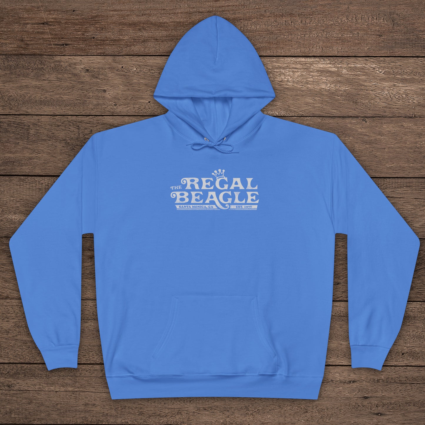 Carolina Blue Hanes EcoSmart pullover hoodie with vintage distressed Regal Beagle logo from Three’s Company on chest, shown on wood floor.