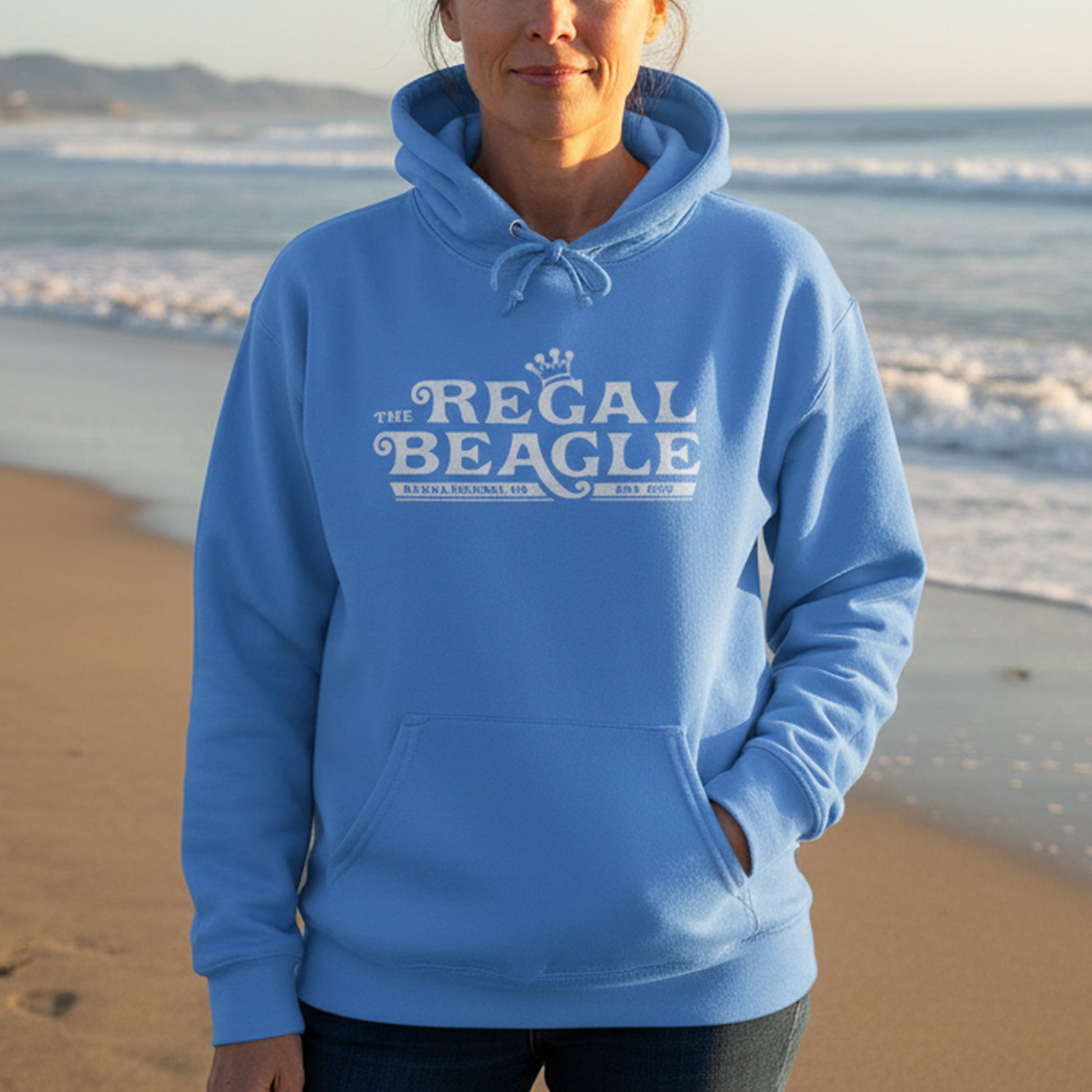 Woman wearing Hanes EcoSmart pullover hoodie with vintage distressed Regal Beagle logo from Three’s Company on chest, standing at a beach.