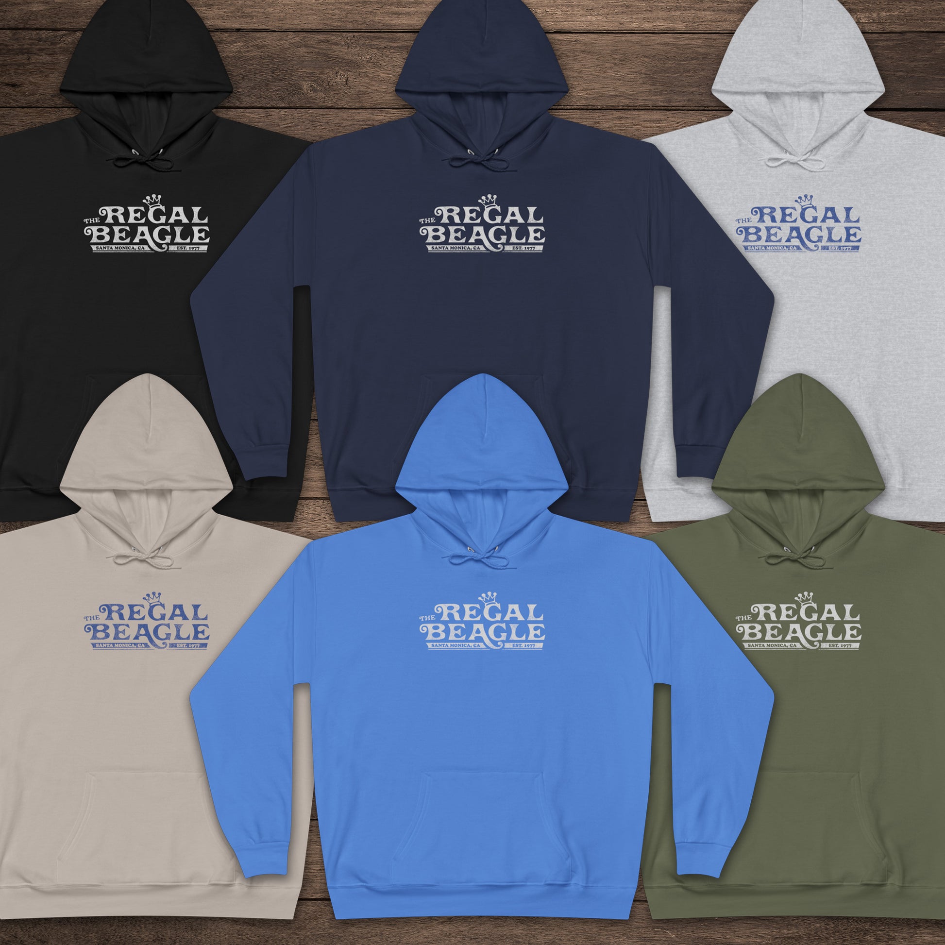 Six Hanes EcoSmart pullover hoodies with vintage distressed Regal Beagle logo from Three’s Company on chest, shown on wood floor.