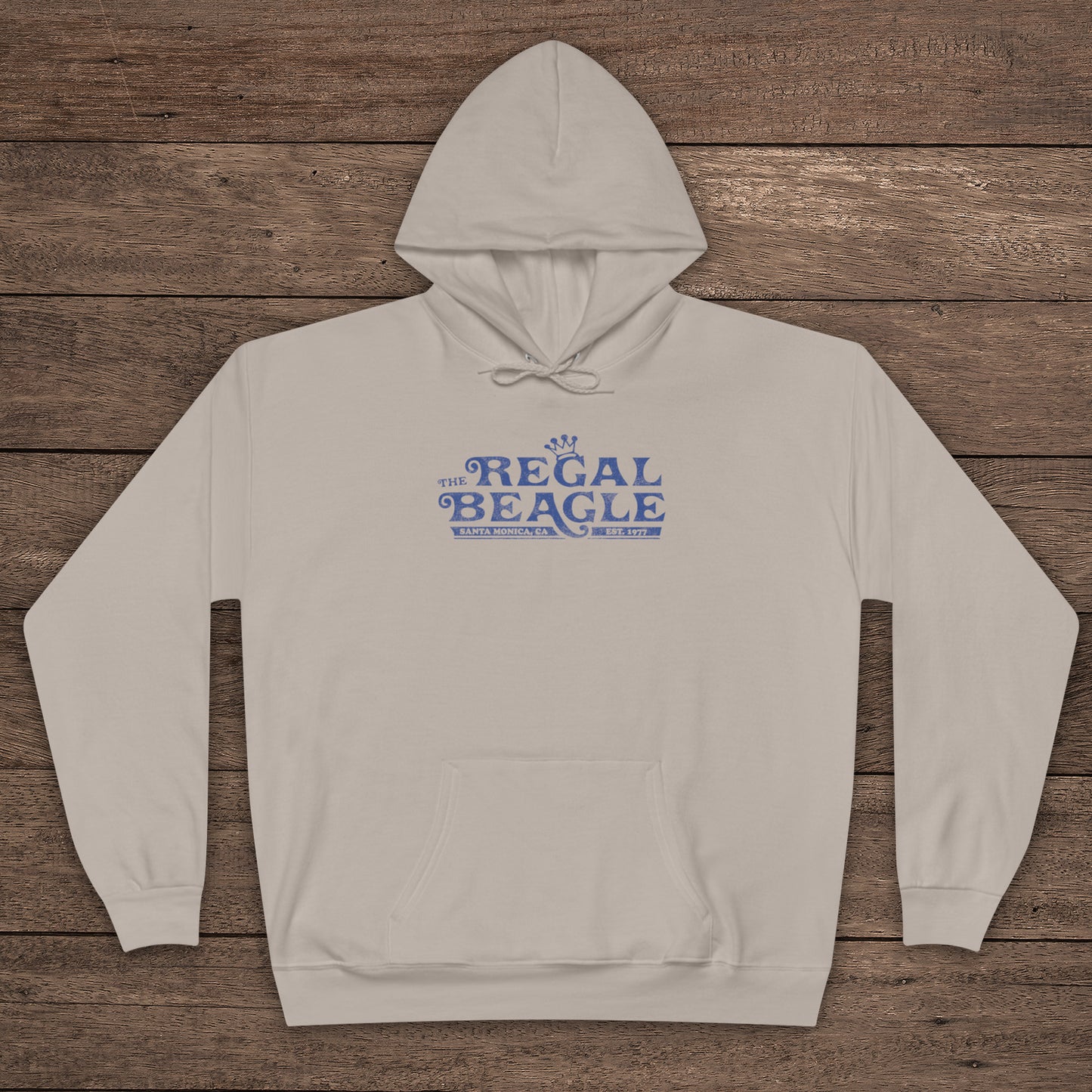 Sand tan Hanes EcoSmart pullover hoodie with vintage distressed Regal Beagle logo from Three’s Company on chest, shown on wood floor.