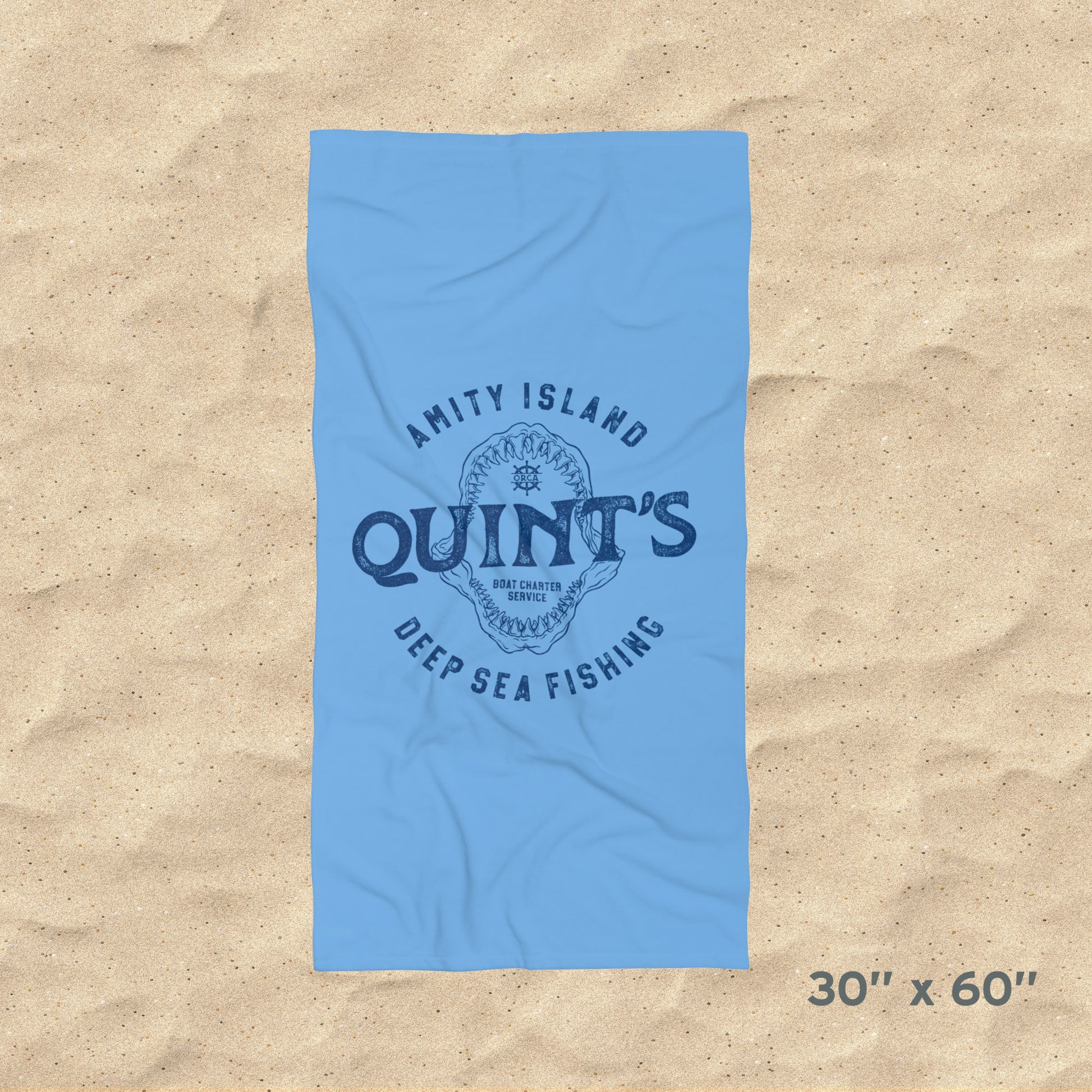 Overhead view of 30x60 Blue beach towel with Quint’s Deep Sea Fishing logo, vintage distressed Jaws inspired design, shown on beach sand.