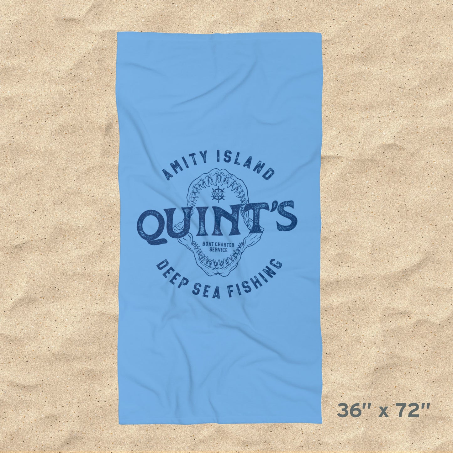 Overhead view of 36x72 Blue beach towel with Quint’s Deep Sea Fishing logo, vintage distressed Jaws inspired design, shown on beach sand.