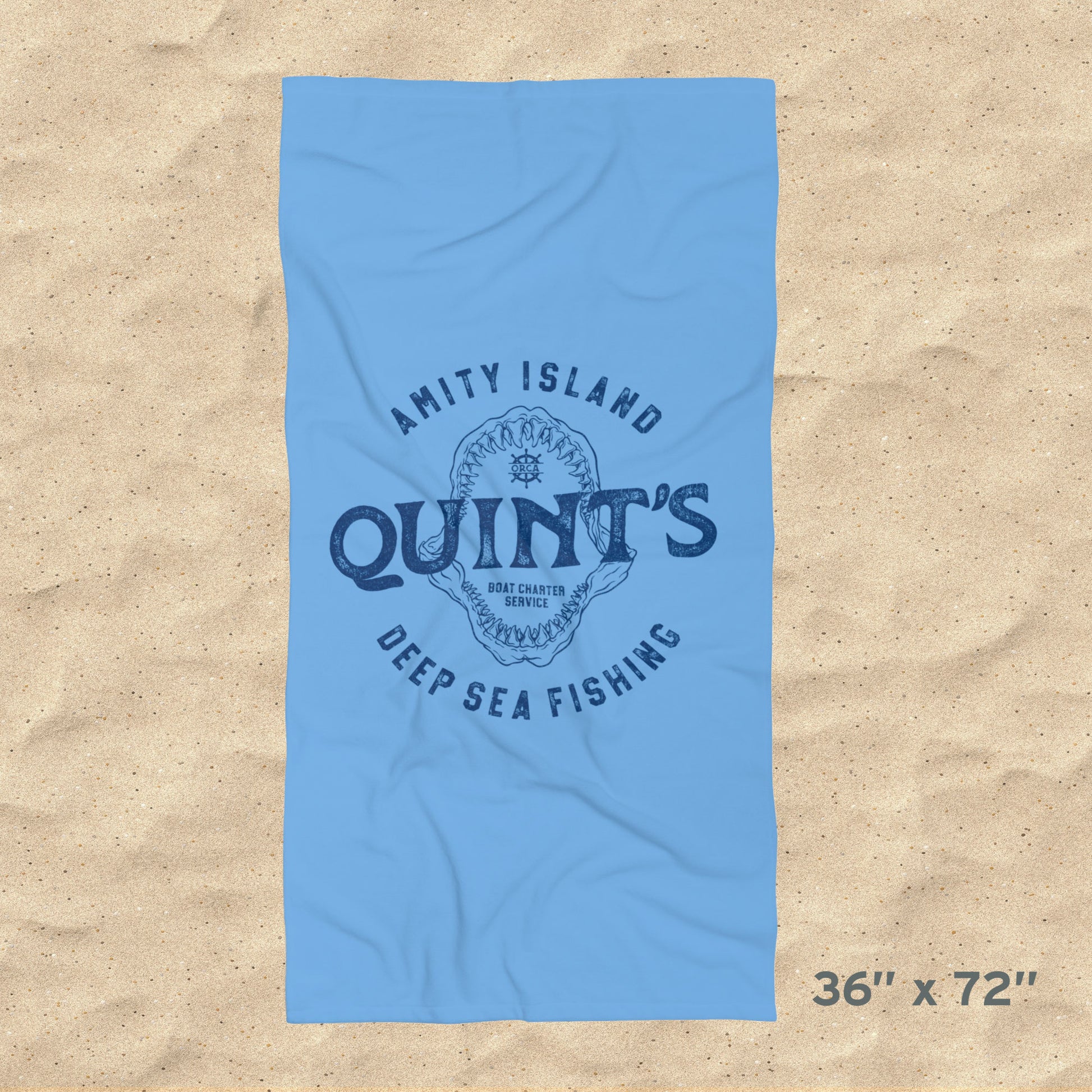 Overhead view of 36x72 Blue beach towel with Quint’s Deep Sea Fishing logo, vintage distressed Jaws inspired design, shown on beach sand.