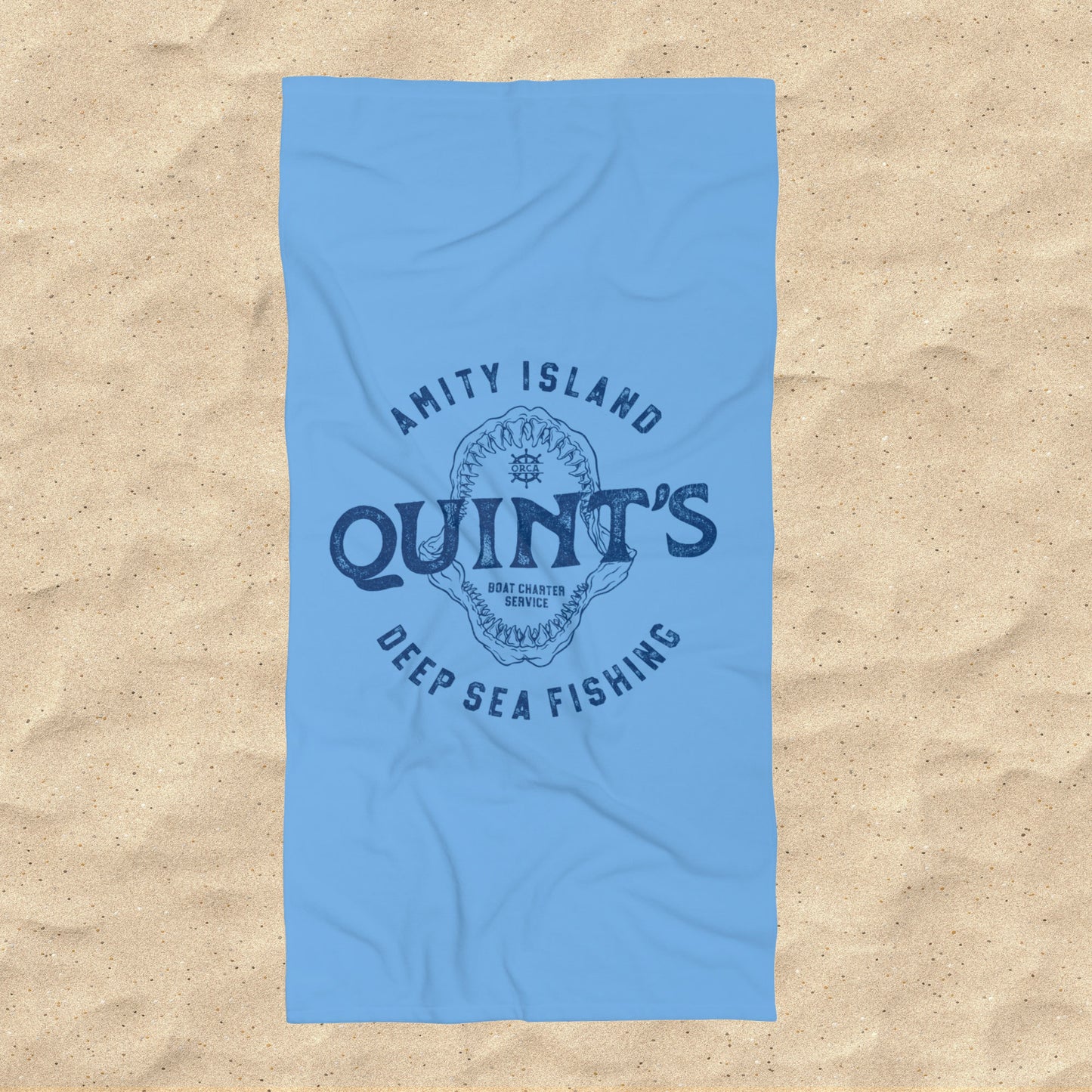 Overhead view of Blue beach towel with Quint’s Deep Sea Fishing logo, vintage distressed Jaws inspired design, shown on beach sand.