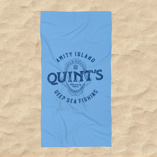 Overhead view of Blue beach towel with Quint’s Deep Sea Fishing logo, vintage distressed Jaws inspired design, shown on beach sand.