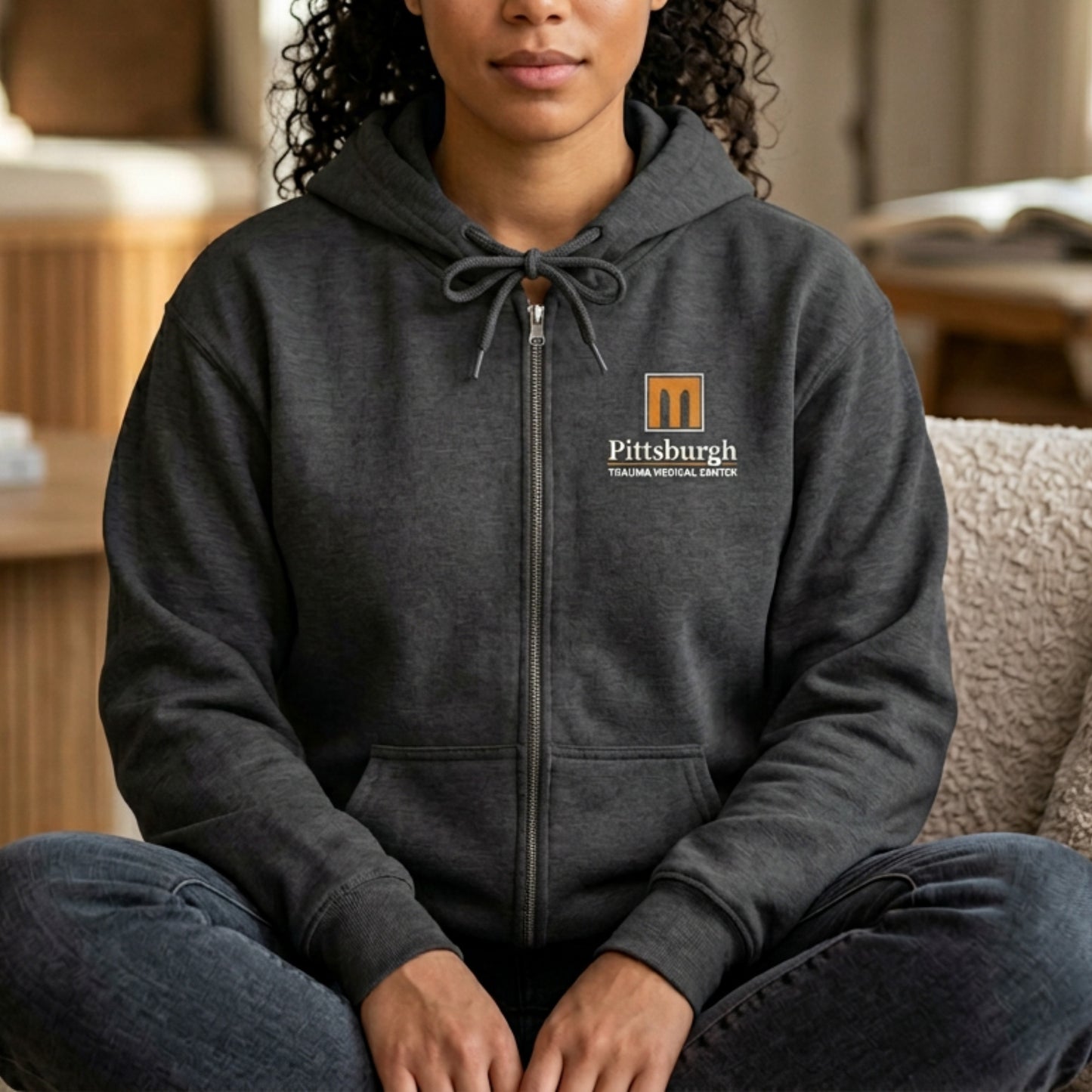 Woman wearing a gray full-zip hoodie with the Pittsburgh Trauma Medical Center (PTMC) logo from The Pitt, printed on the left chest