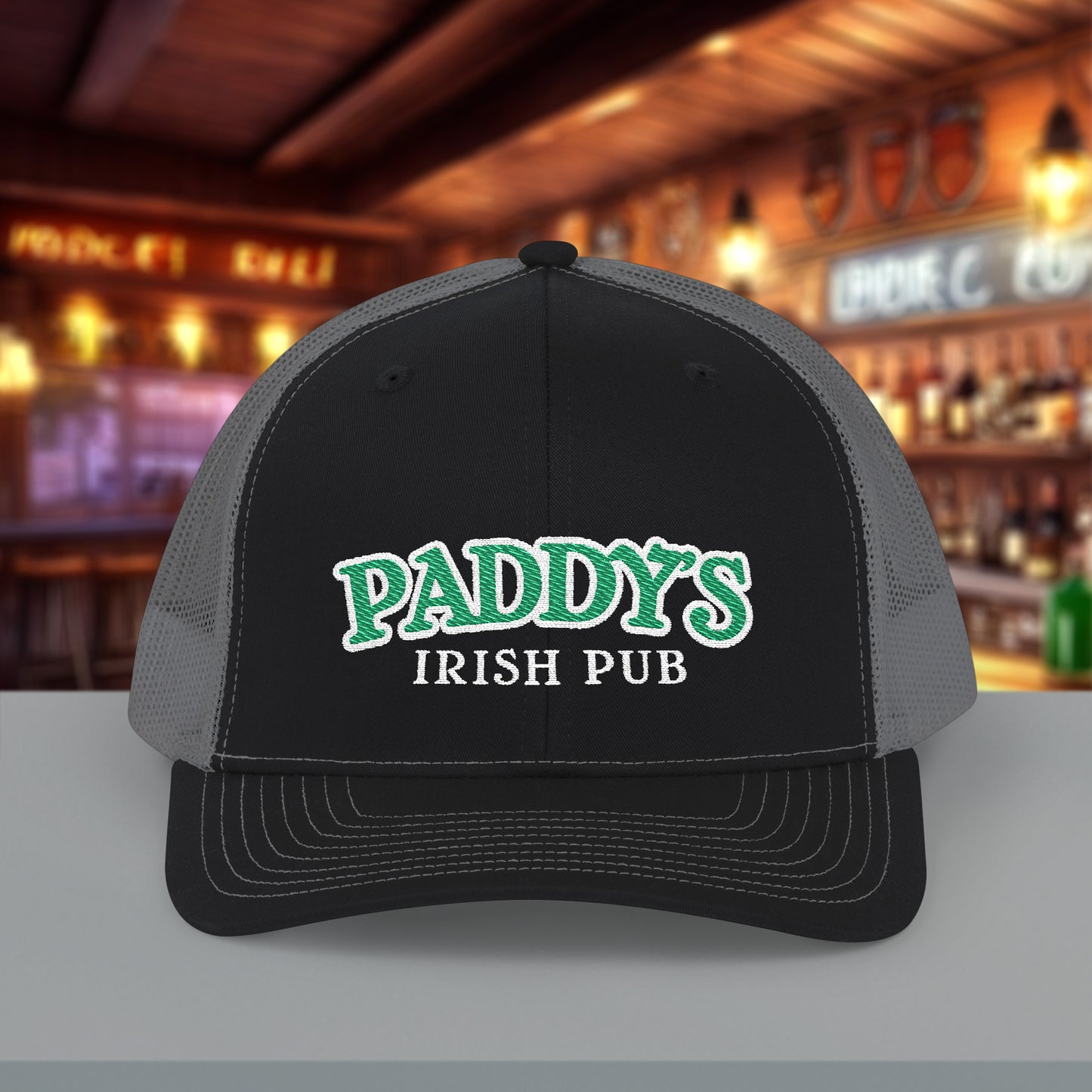 Front view of Richardson 112 black charcoal trucker cap with green Paddy’s Irish Pub embroidered logo shown in a bar.