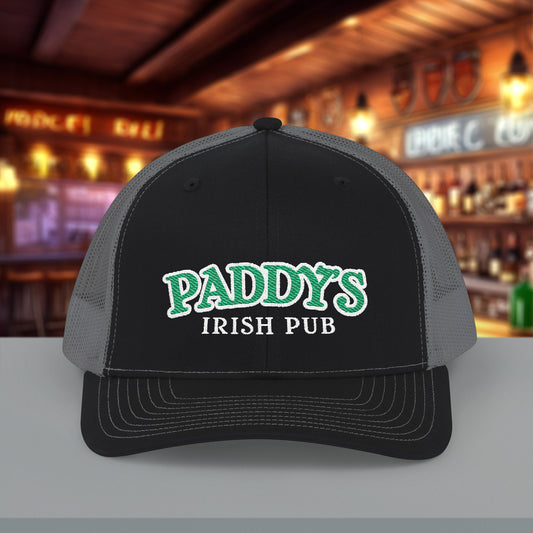 Front view of Richardson 112 black charcoal trucker cap with green Paddy’s Irish Pub embroidered logo shown in a bar.