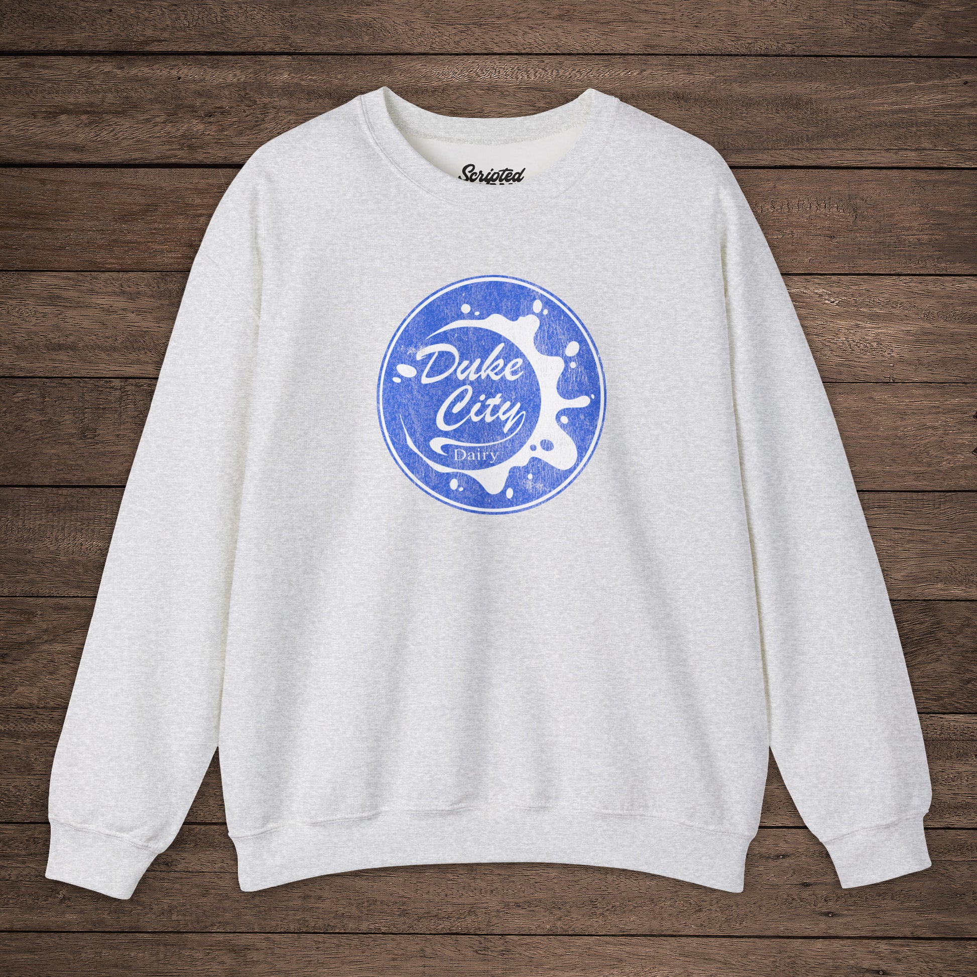Ash sweatshirt with blue vintage distressed Duke City Dairy logo from the TV Show Pluribus, displayed flat on wood background.