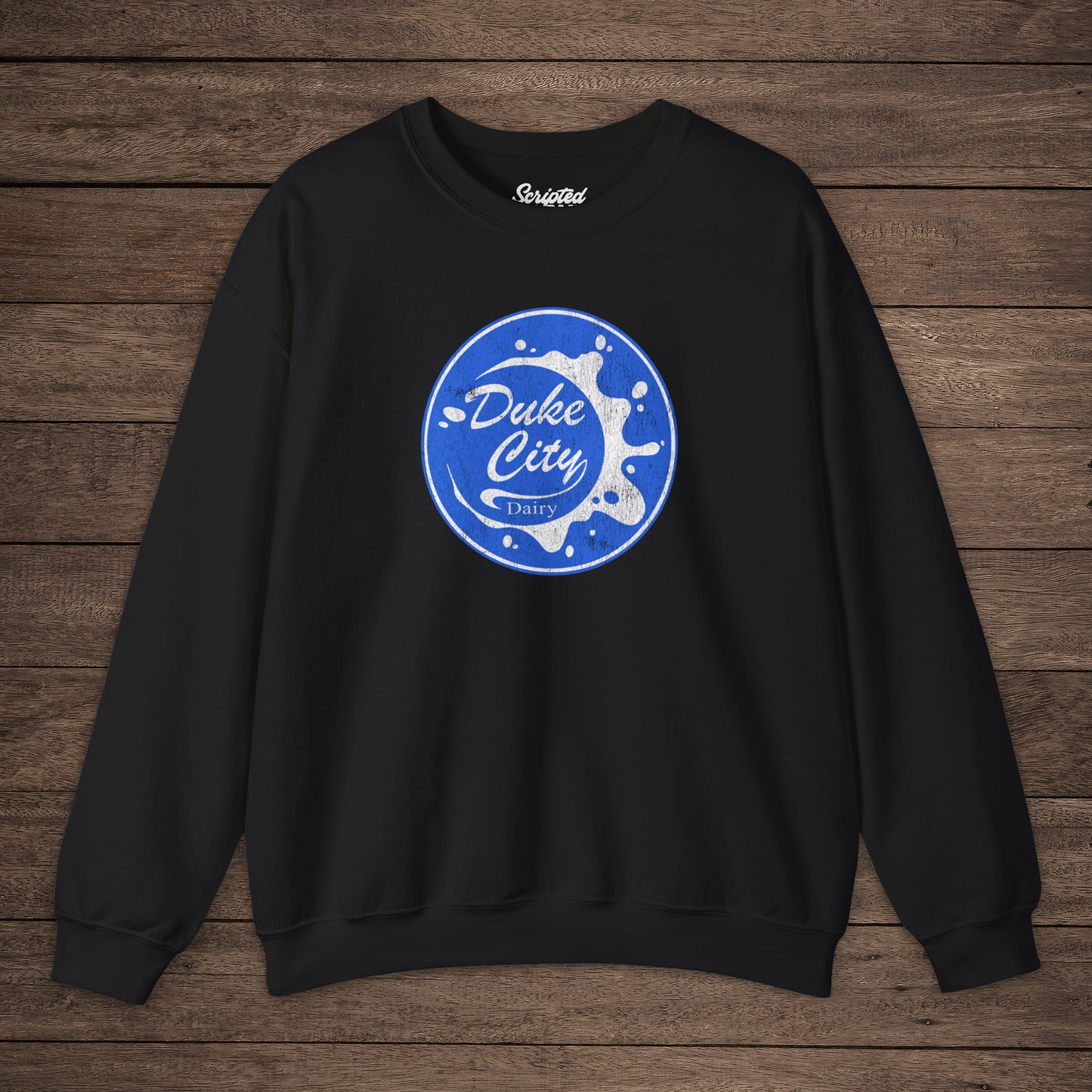 Black sweatshirt with blue vintage distressed Duke City Dairy logo from the TV Show Pluribus, displayed flat on wood background.