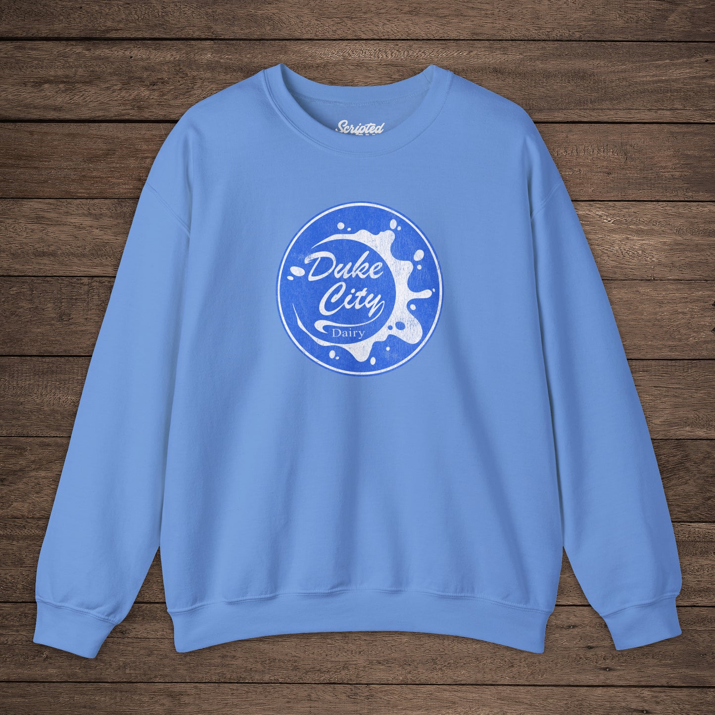 Light blue sweatshirt with blue vintage distressed Duke City Dairy logo from the TV Show Pluribus, displayed flat on wood background.