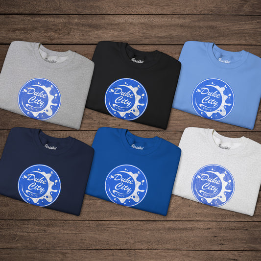 Six folded sweatshirts with blue vintage distressed Duke City Dairy logo from the TV Show Pluribus, displayed flat on wood background.