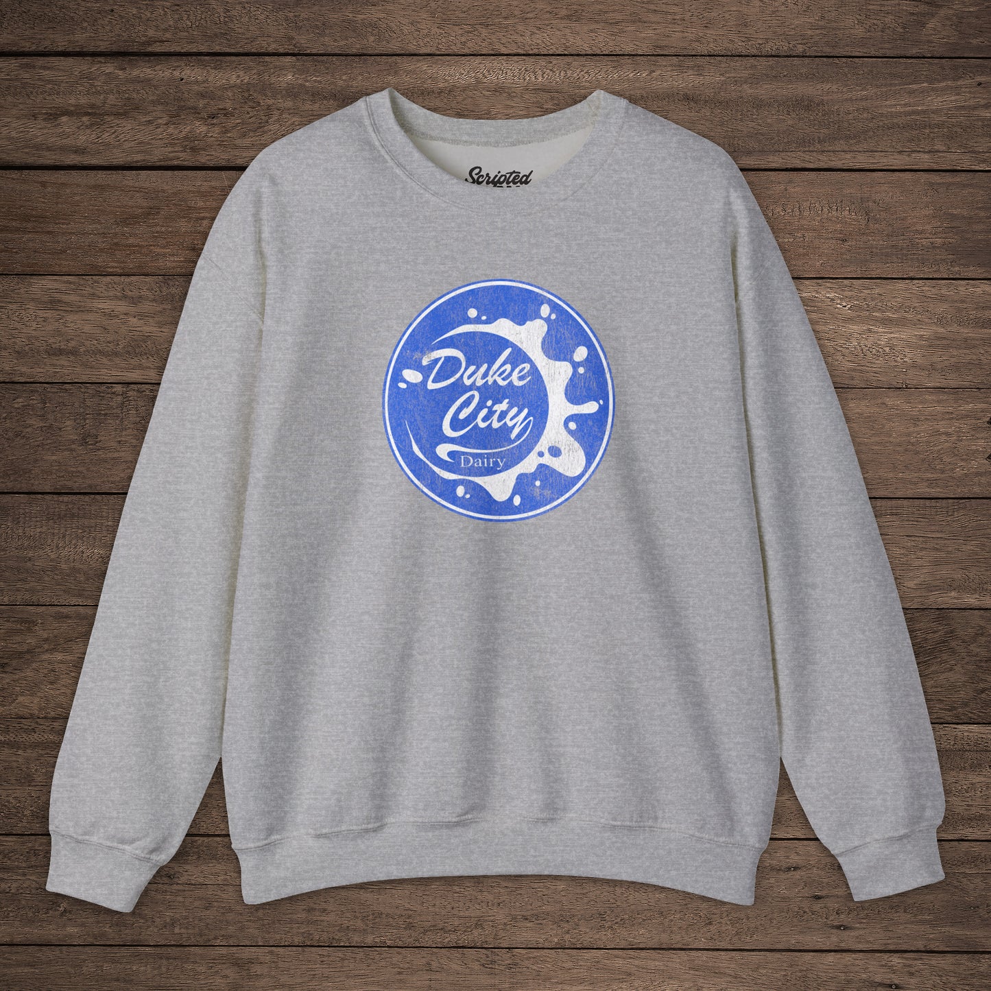 Heather sport gray sweatshirt with blue vintage distressed Duke City Dairy logo from the TV Show Pluribus, displayed flat on wood background.