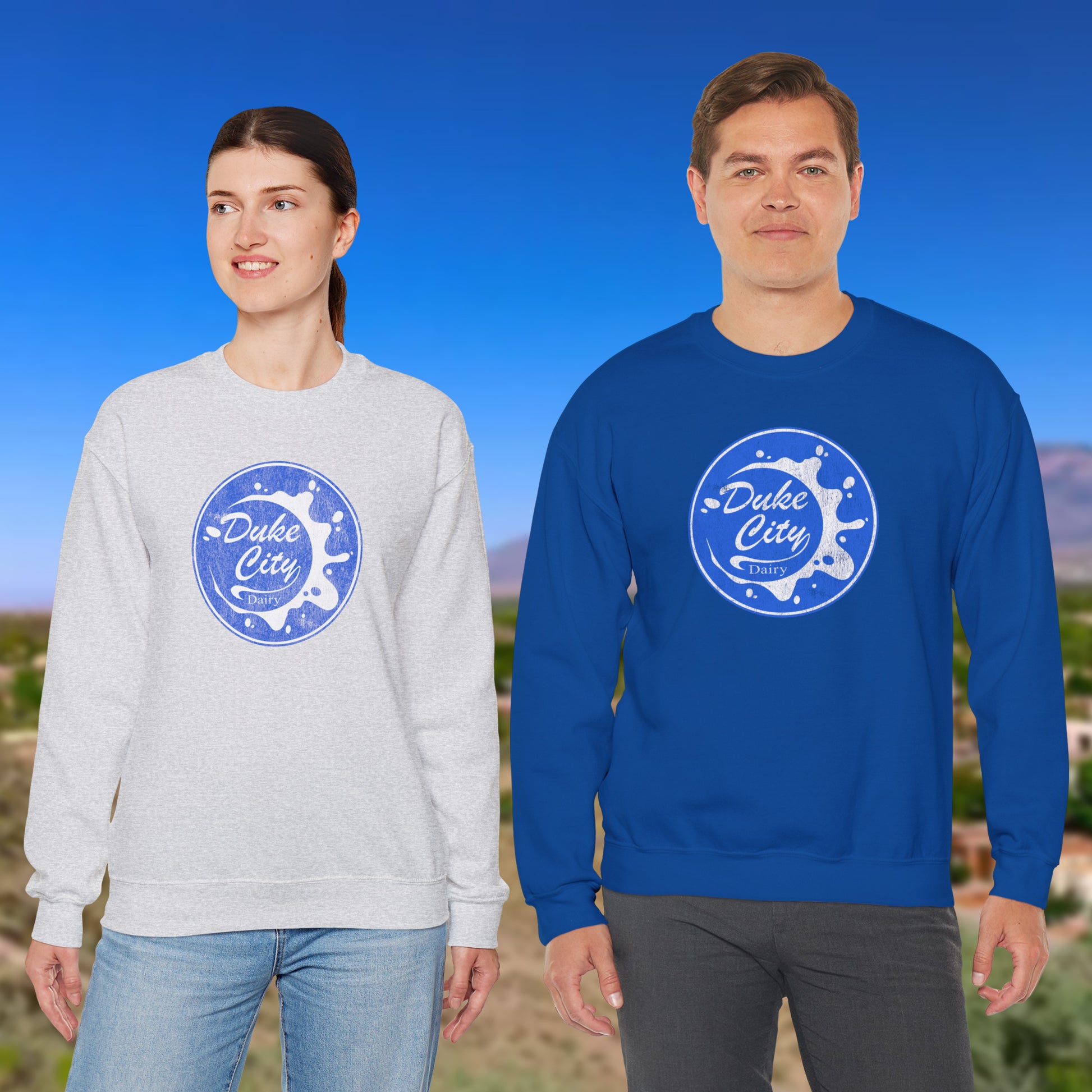 Two people standing outside wearing sweatshirts with blue vintage distressed Duke City Dairy logo from the TV Show Pluribus.
