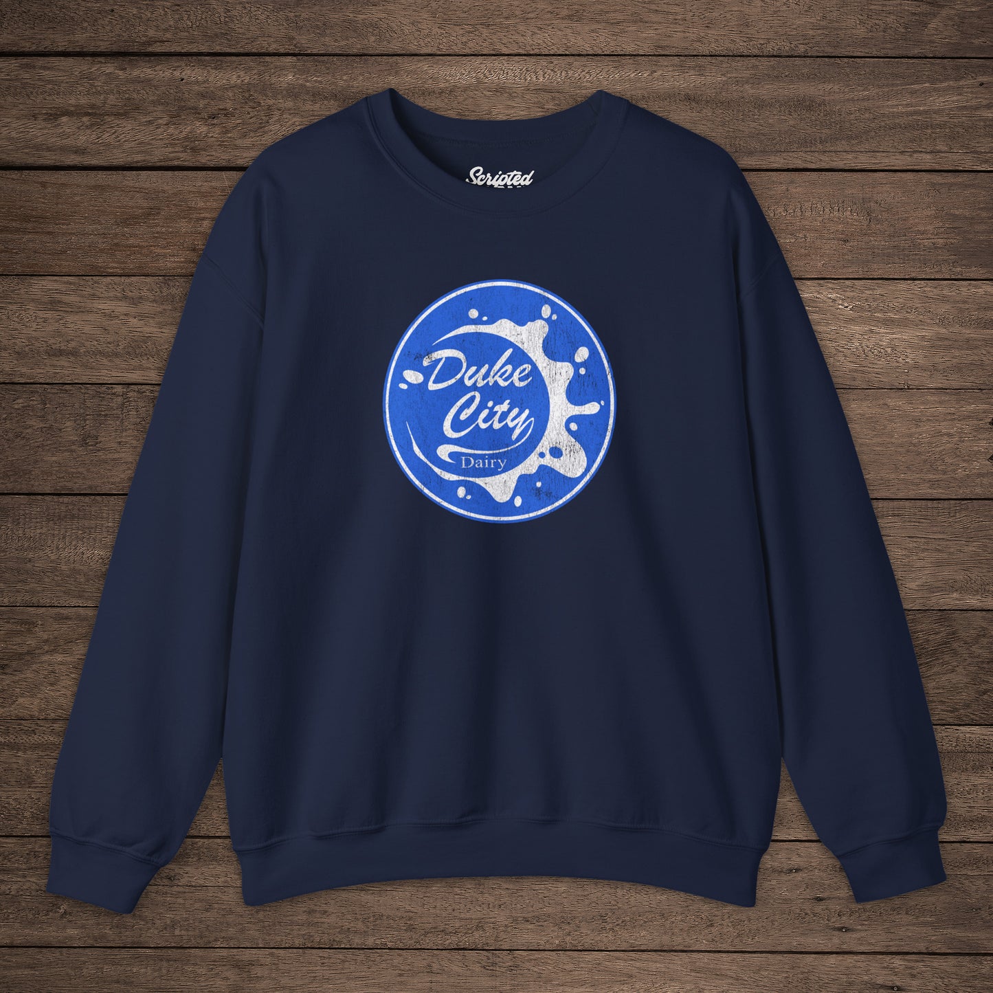 Navy blue sweatshirt with blue vintage distressed Duke City Dairy logo from the TV Show Pluribus, displayed flat on wood background.
