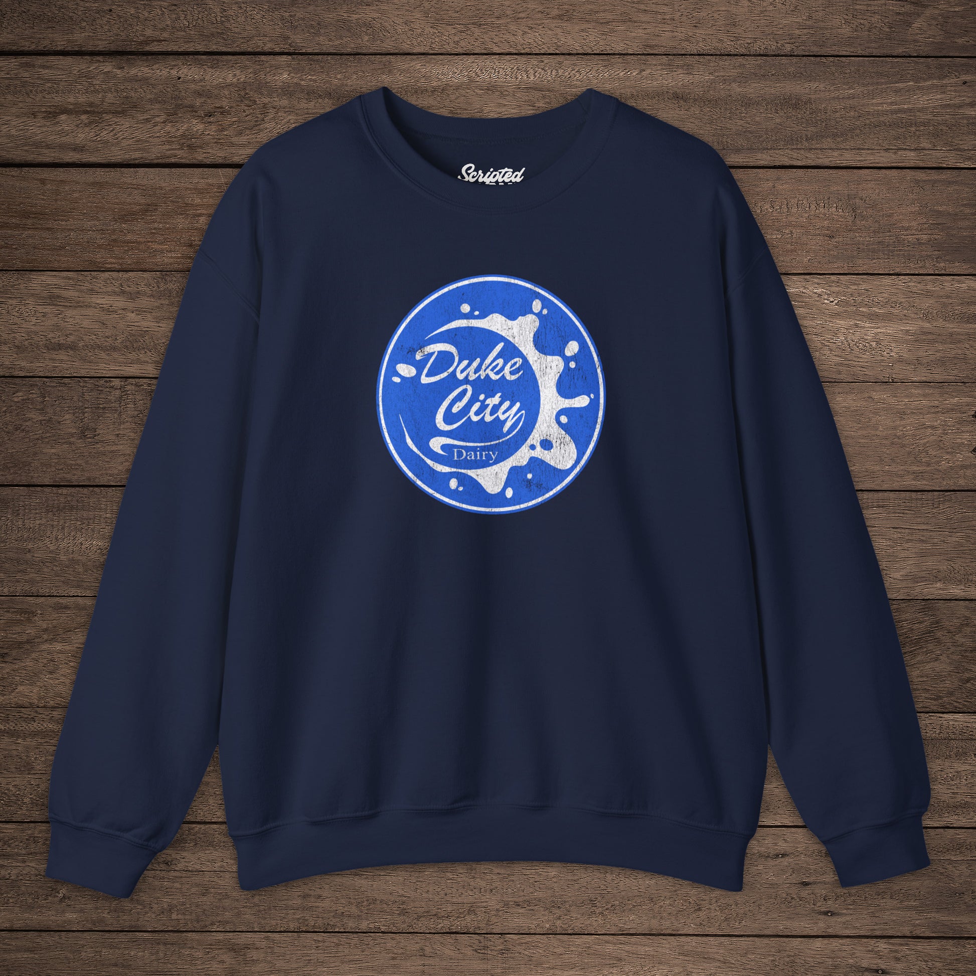 Navy blue sweatshirt with blue vintage distressed Duke City Dairy logo from the TV Show Pluribus, displayed flat on wood background.