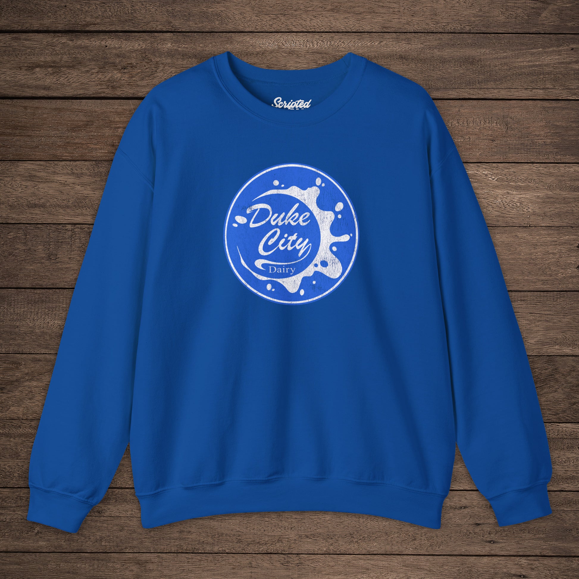 Royal blue sweatshirt with blue vintage distressed Duke City Dairy logo from the TV Show Pluribus, displayed flat on wood background.