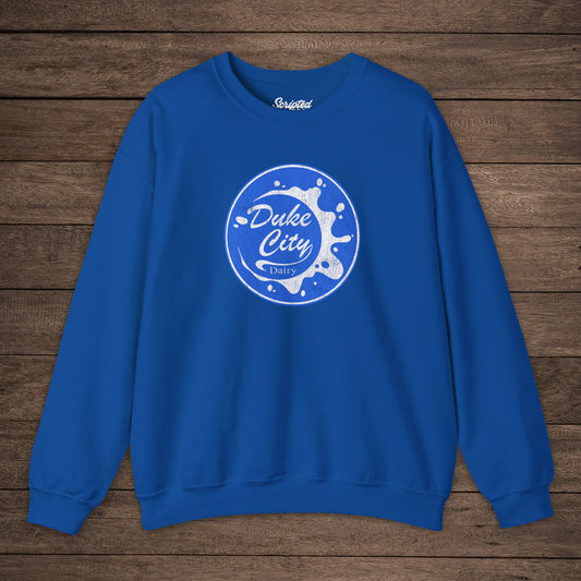 Royal blue sweatshirt with blue vintage distressed Duke City Dairy logo from the TV Show Pluribus, displayed flat on wood background.