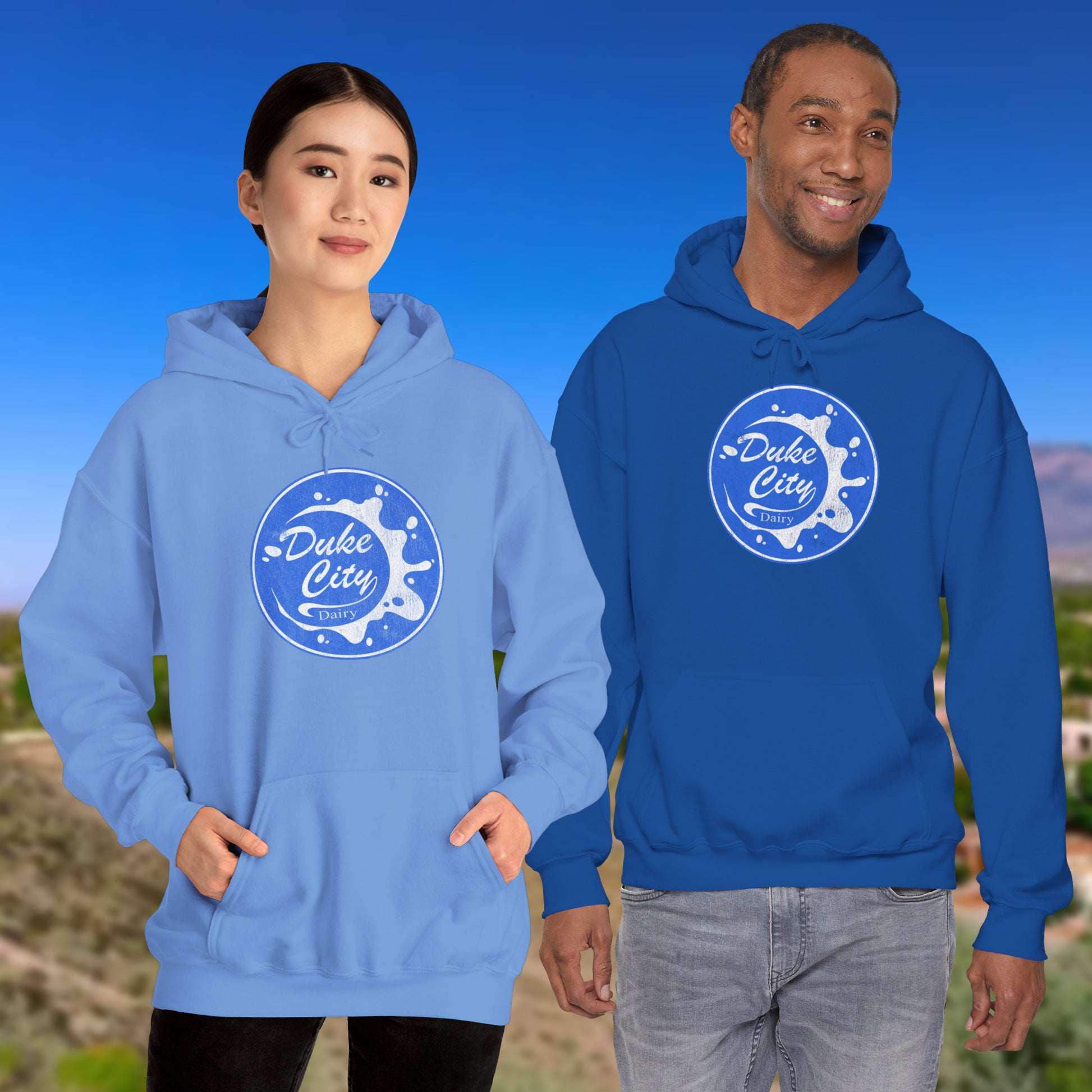 Two people wearing blue hoodies with 'Duke City' logo from Pluribus against a scenic background