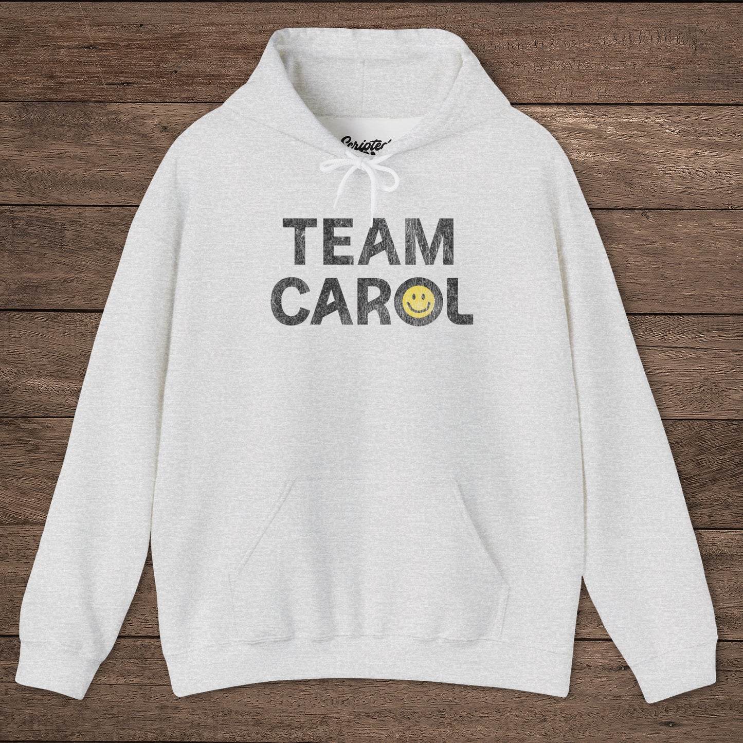 Ash heather unisex Heavy Blend pullover hoodie sweatshirt with TEAM CAROL vintage distressed smiley graphic, on wood surface
