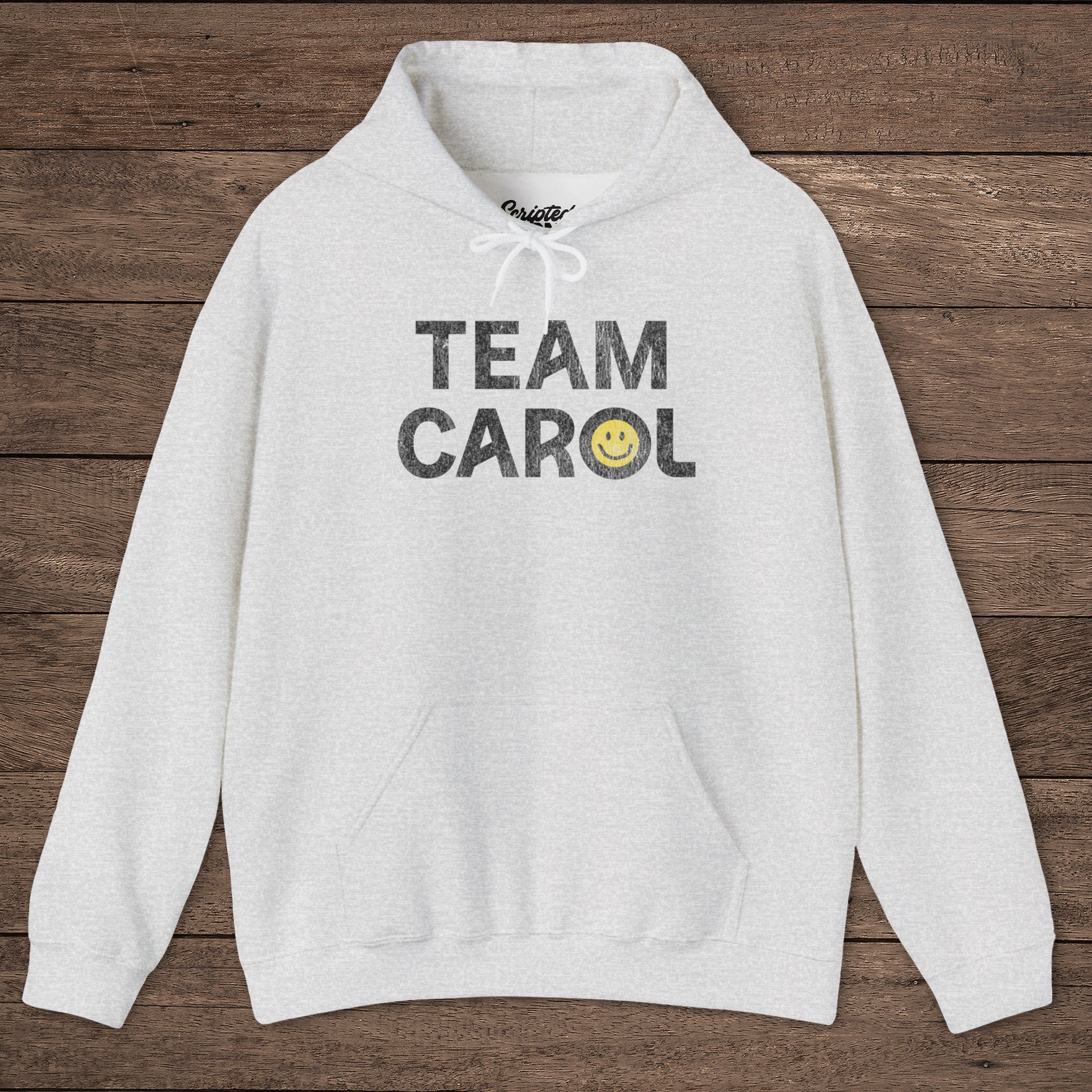 Ash heather unisex Heavy Blend pullover hoodie sweatshirt with TEAM CAROL vintage distressed smiley graphic, on wood surface