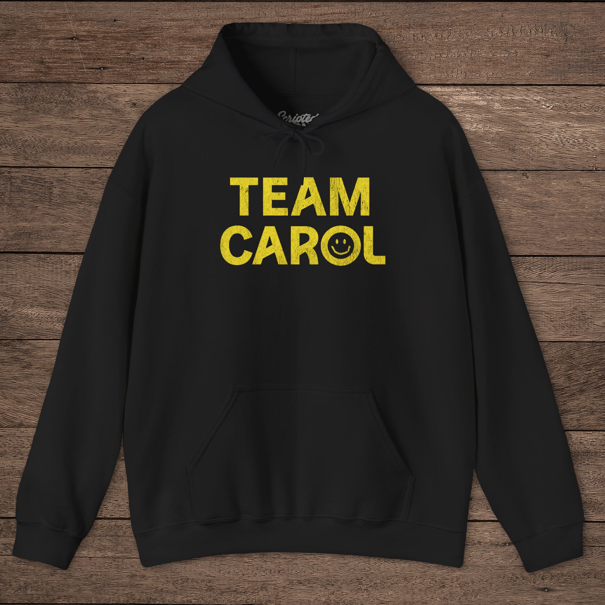 Black unisex Heavy Blend pullover hoodie sweatshirt with TEAM CAROL vintage distressed smiley graphic, on wood surface