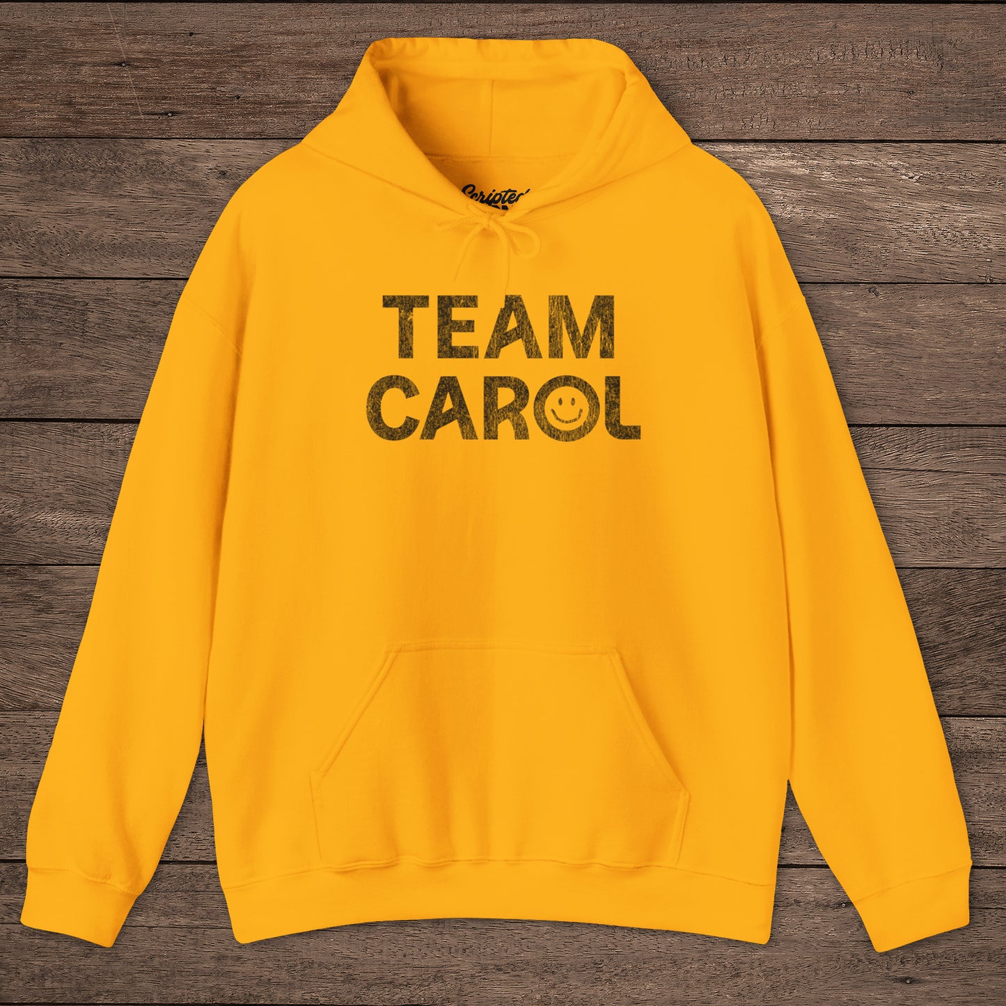 Gold yellow unisex Heavy Blend pullover hoodie sweatshirt with TEAM CAROL vintage distressed smiley graphic, on wood surface