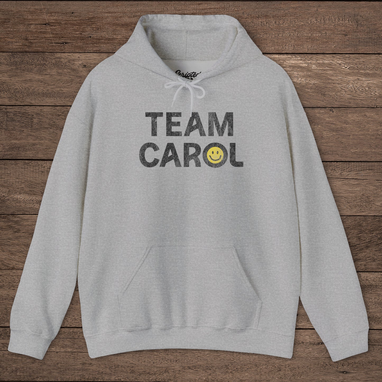 Heather gray unisex Heavy Blend pullover hoodie sweatshirt with TEAM CAROL vintage distressed smiley graphic, on wood surface
