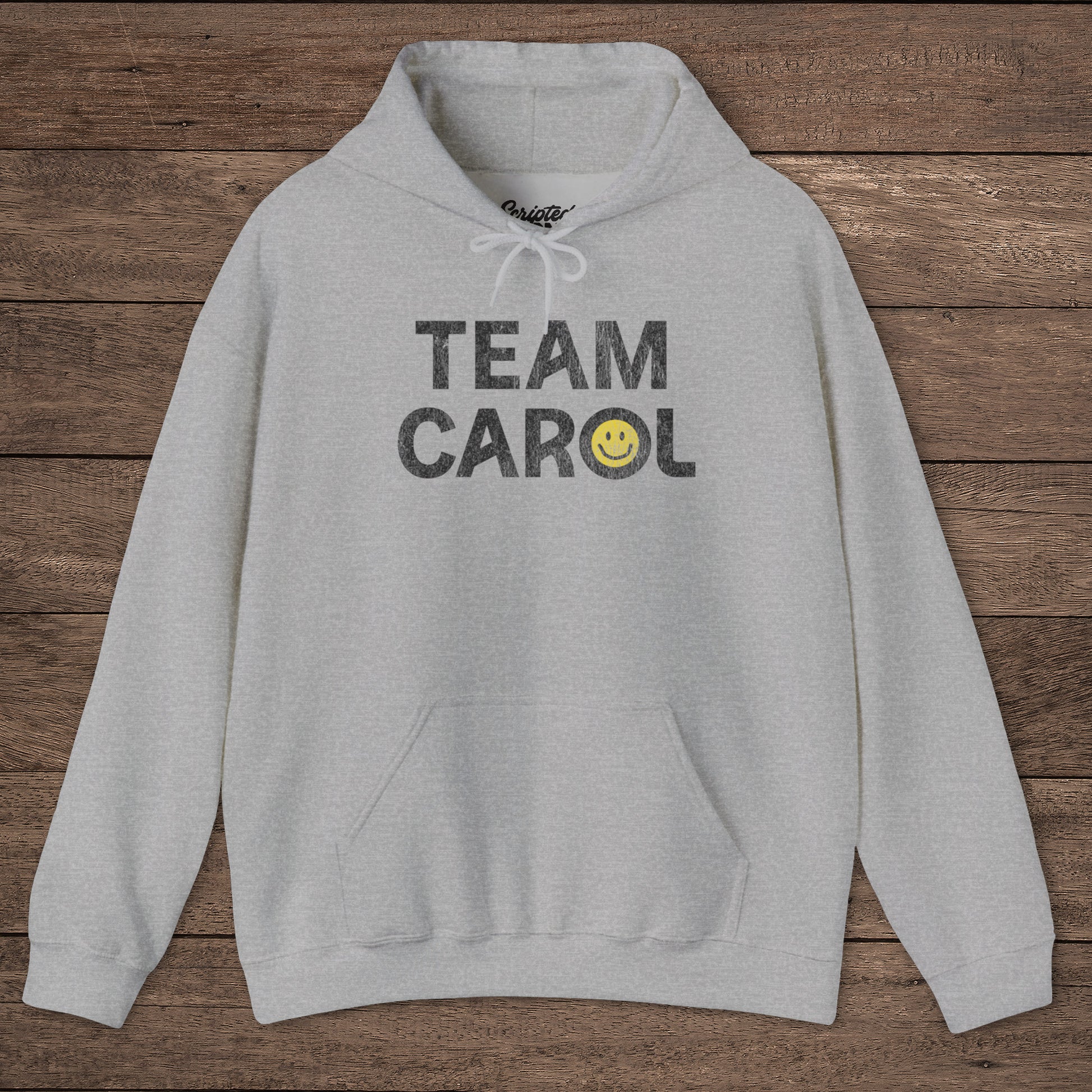 Heather gray unisex Heavy Blend pullover hoodie sweatshirt with TEAM CAROL vintage distressed smiley graphic, on wood surface