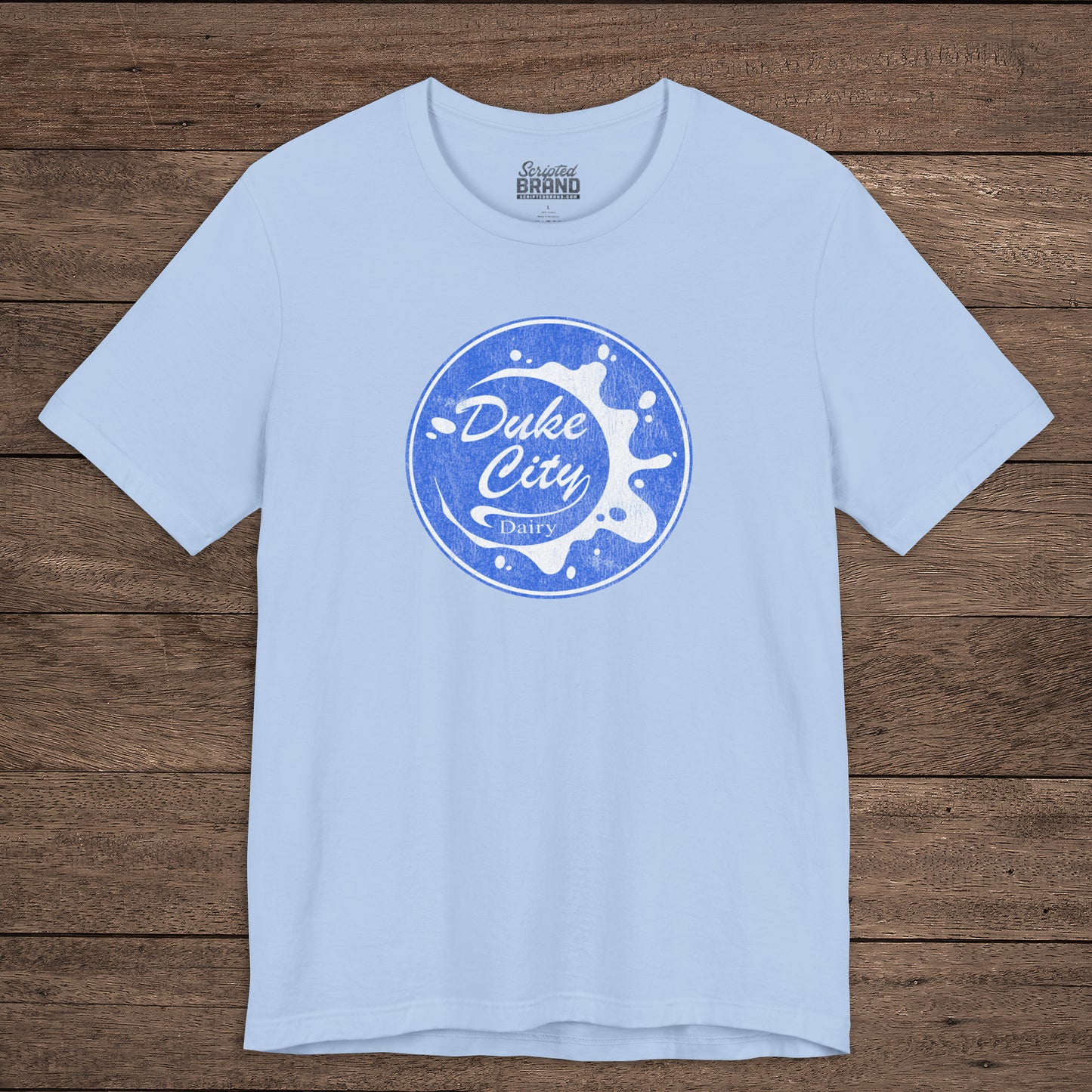 Light blue t-shirt with vintage distressed Duke City Dairy logo inspired by TV Show Pluribus, displayed flat wood background.