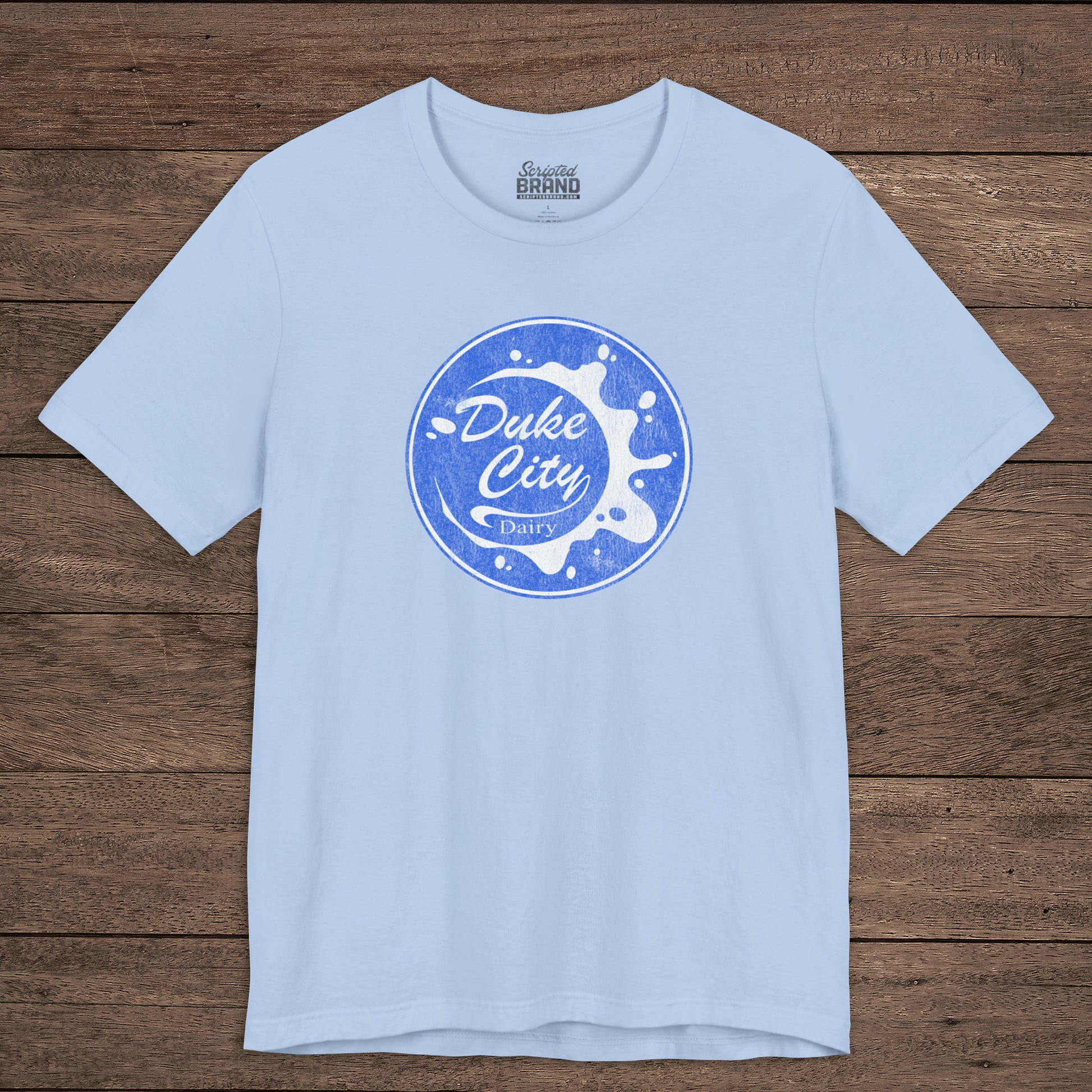 Light blue t-shirt with vintage distressed Duke City Dairy logo inspired by TV Show Pluribus, displayed flat wood background.