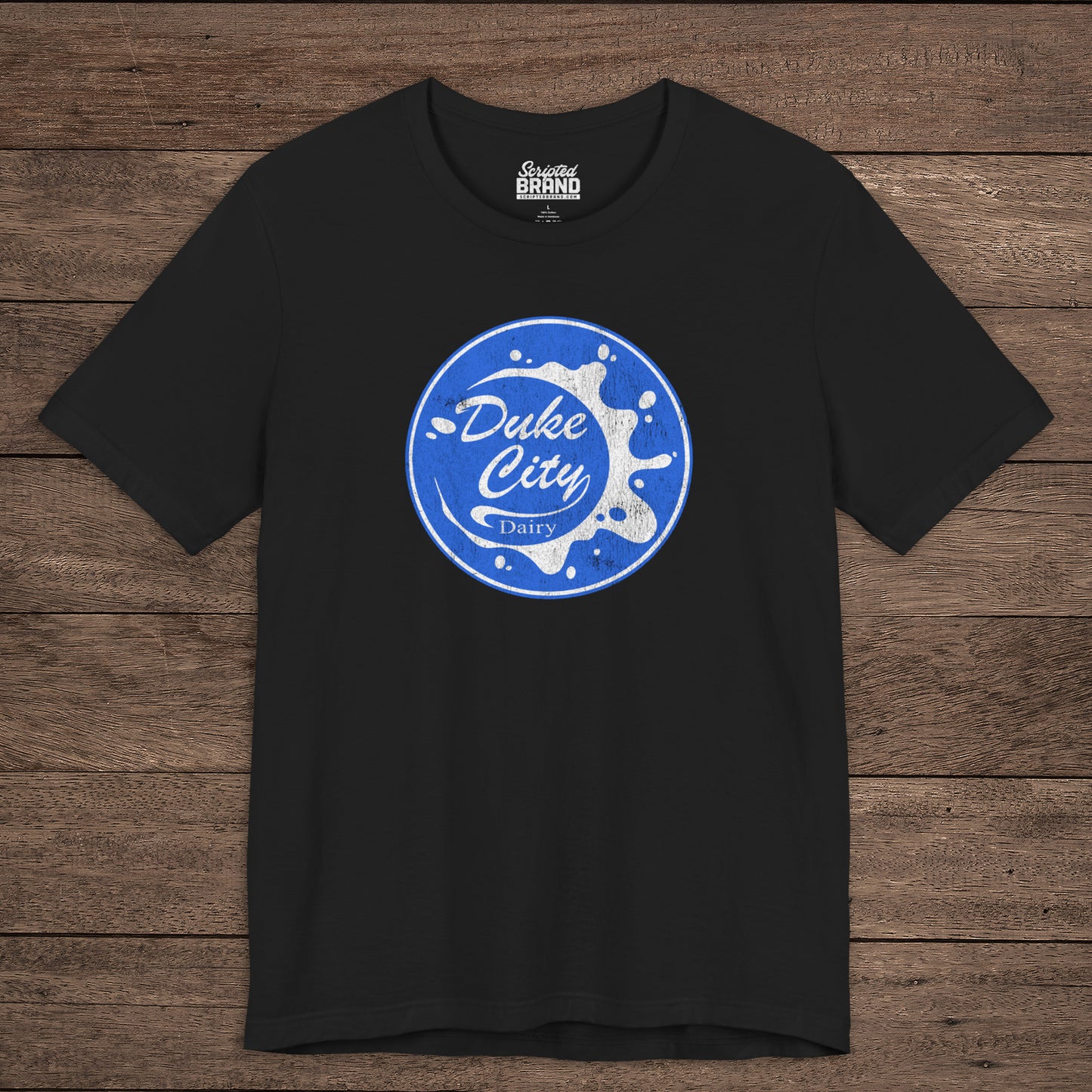 Black t-shirt with vintage distressed Duke City Dairy logo inspired by TV Show Pluribus, displayed flat wood background.
