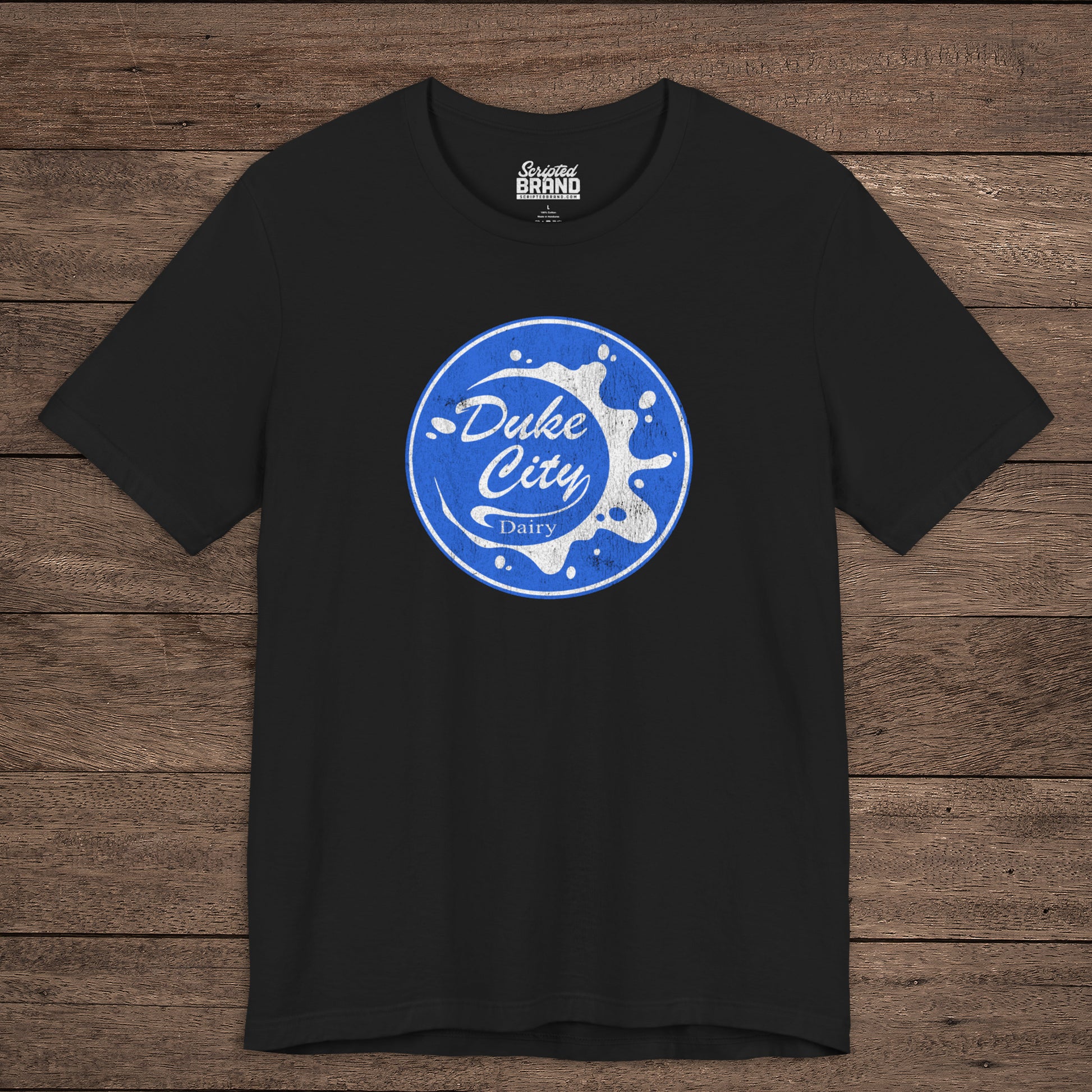 Black t-shirt with vintage distressed Duke City Dairy logo inspired by TV Show Pluribus, displayed flat wood background.
