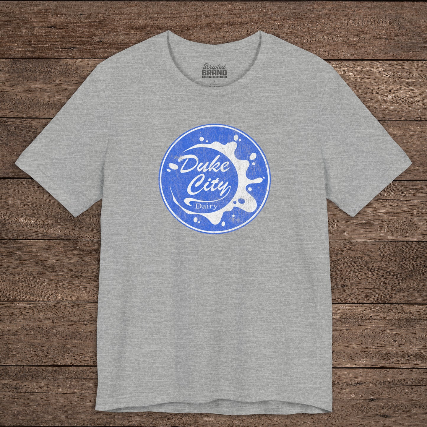 Heather gray t-shirt with vintage distressed Duke City Dairy logo inspired by TV Show Pluribus, displayed flat wood background.