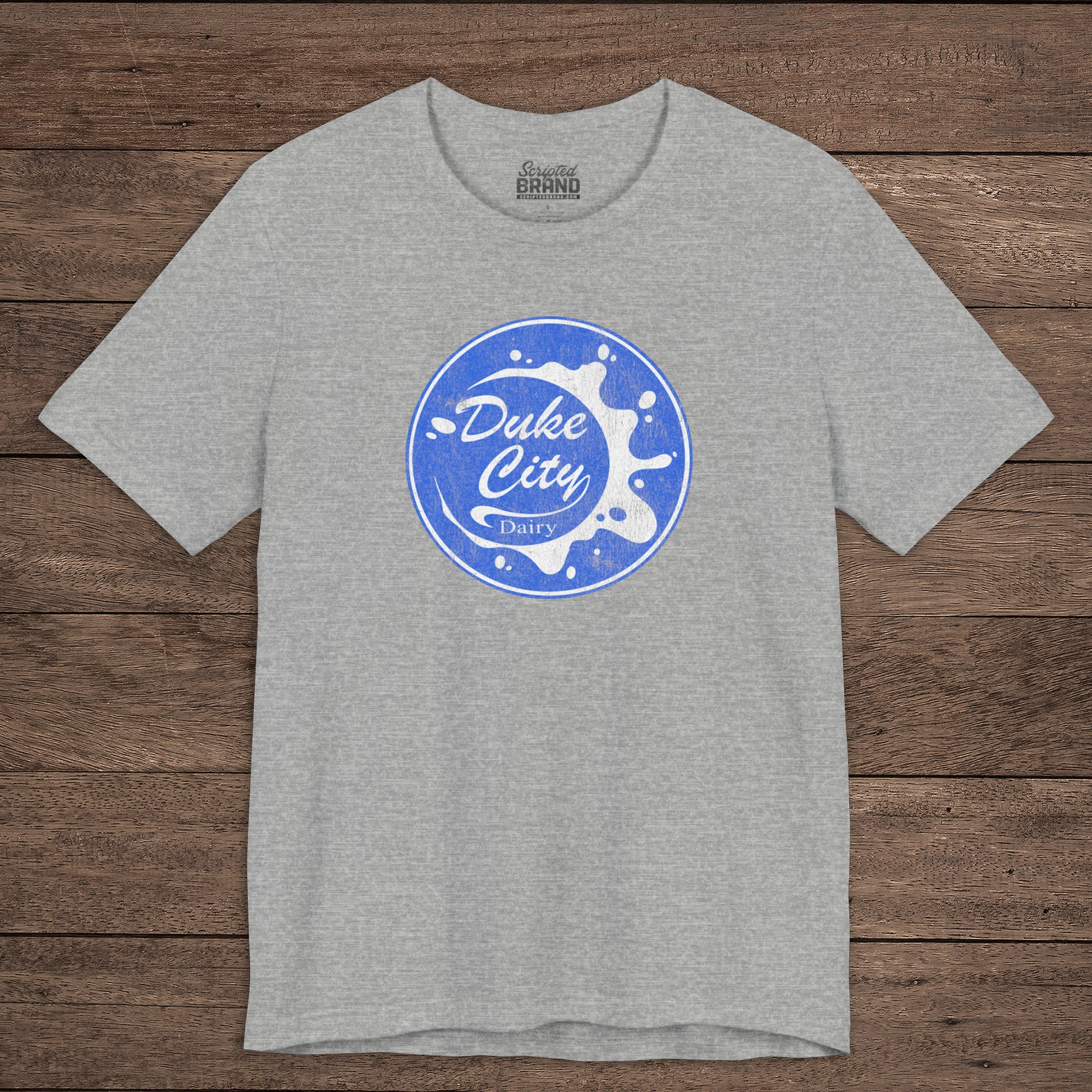Heather gray t-shirt with vintage distressed Duke City Dairy logo inspired by TV Show Pluribus, displayed flat wood background.