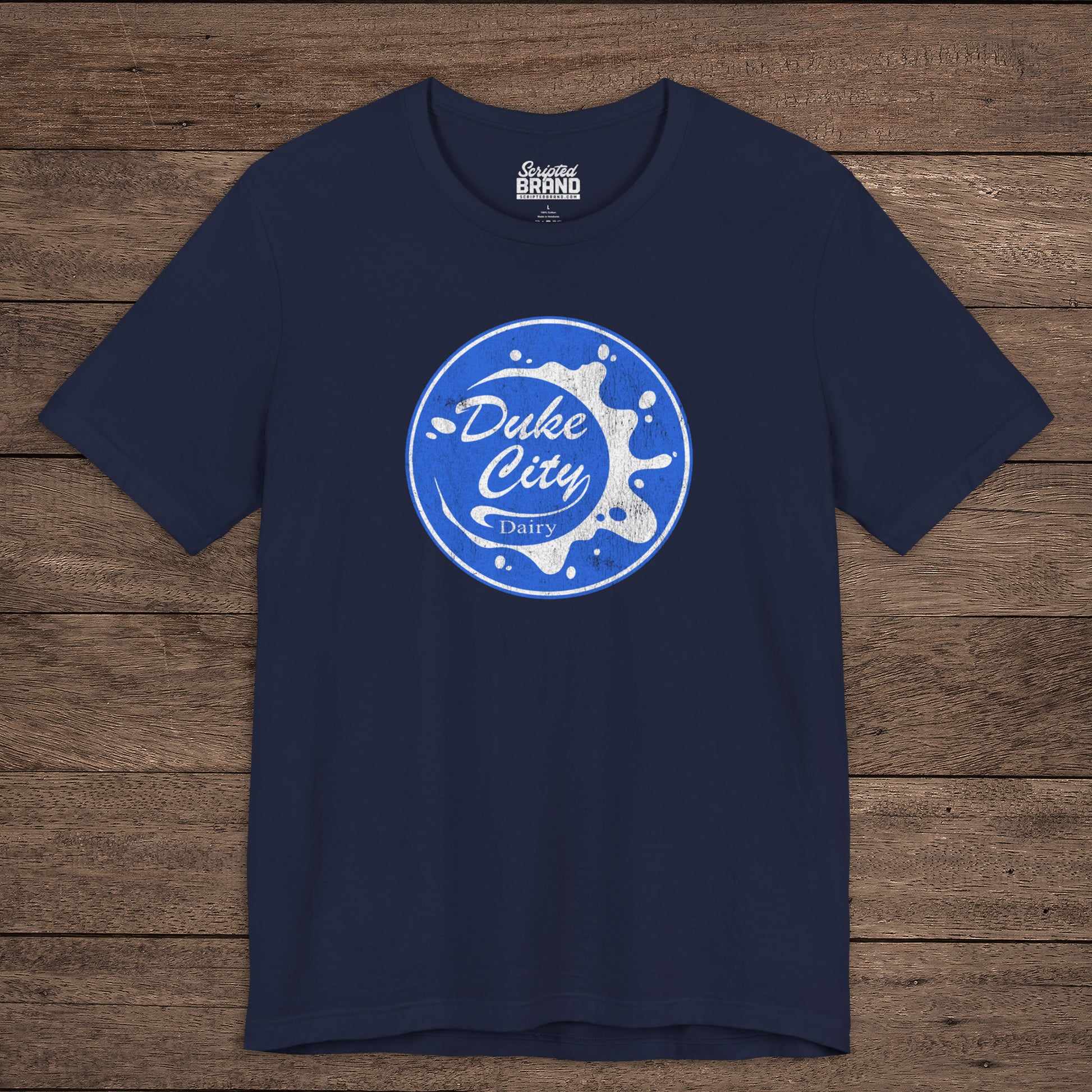 Navy blue t-shirt with vintage distressed Duke City Dairy logo inspired by TV Show Pluribus, displayed flat wood background.