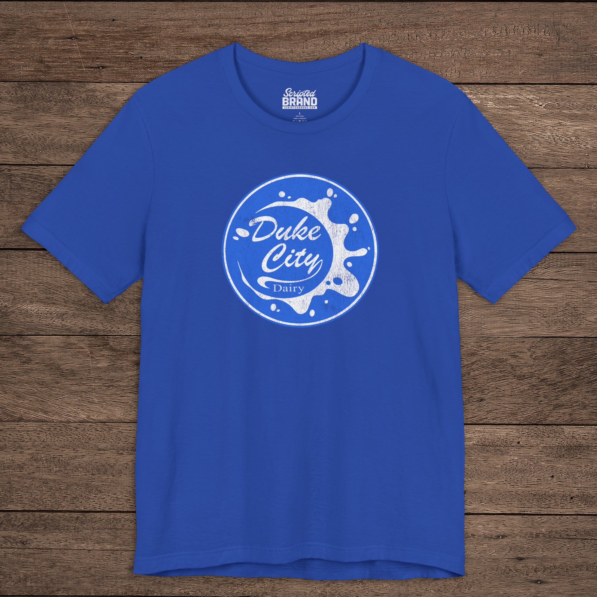 Royal blue t-shirt with vintage distressed Duke City Dairy logo inspired by TV Show Pluribus, displayed flat wood background.