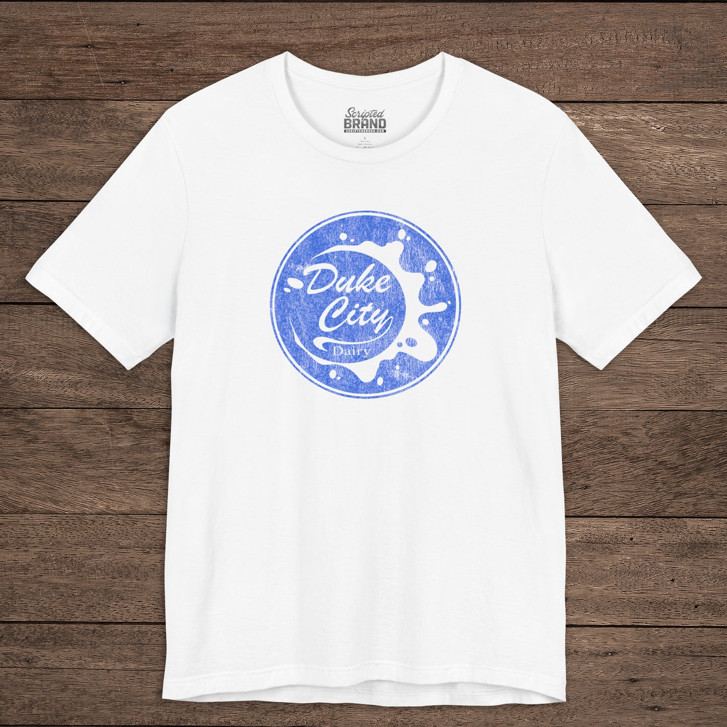 White t-shirt with vintage distressed Duke City Dairy logo inspired by TV Show Pluribus, displayed flat wood background.