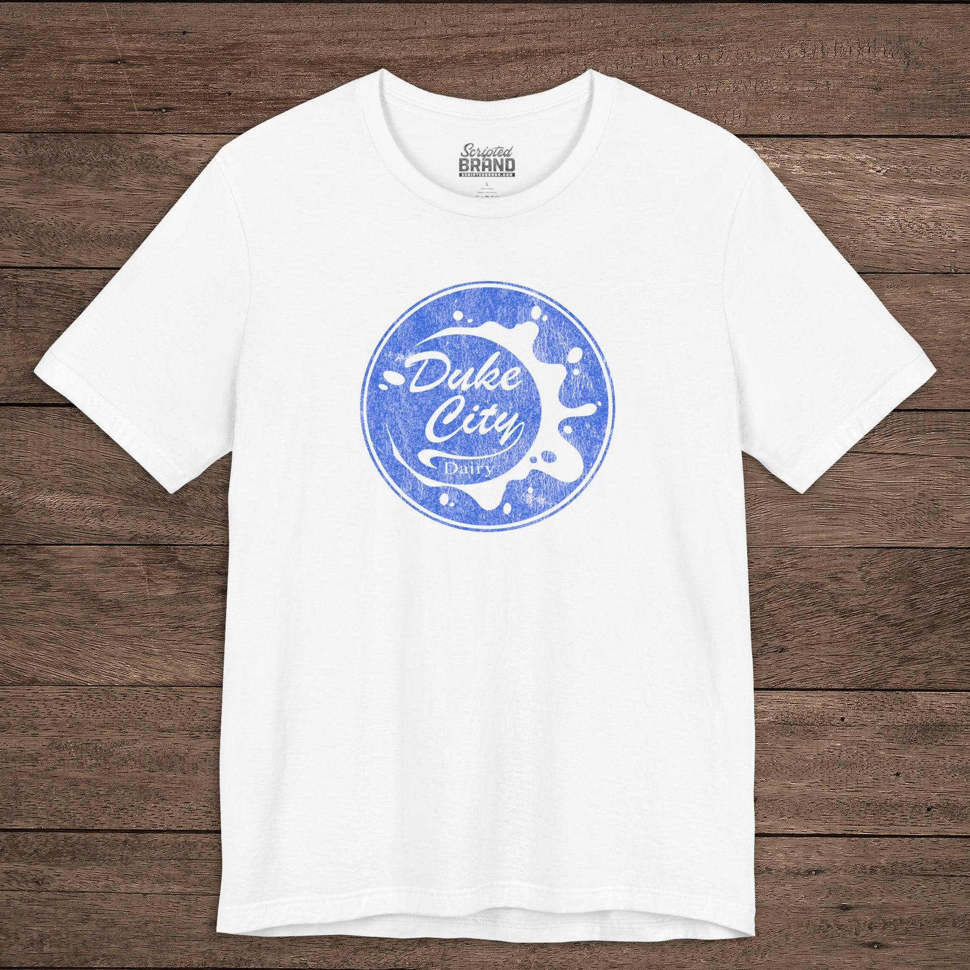 White t-shirt with vintage distressed Duke City Dairy logo inspired by TV Show Pluribus, displayed flat wood background.