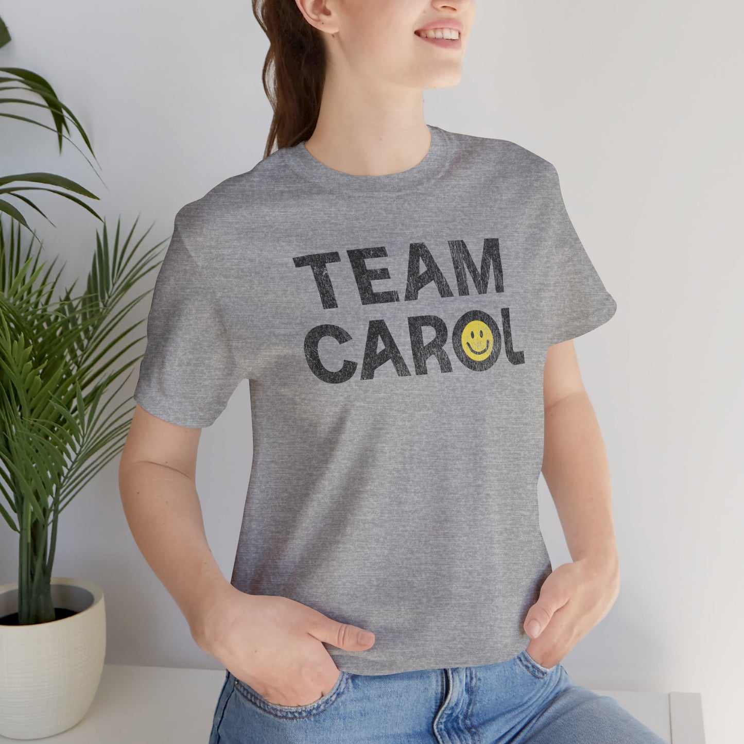 Person wearing heather gray TEAM CAROL t-shirt inspired by Pluribus with vintage distressed smiley face graphic, seated near a plant