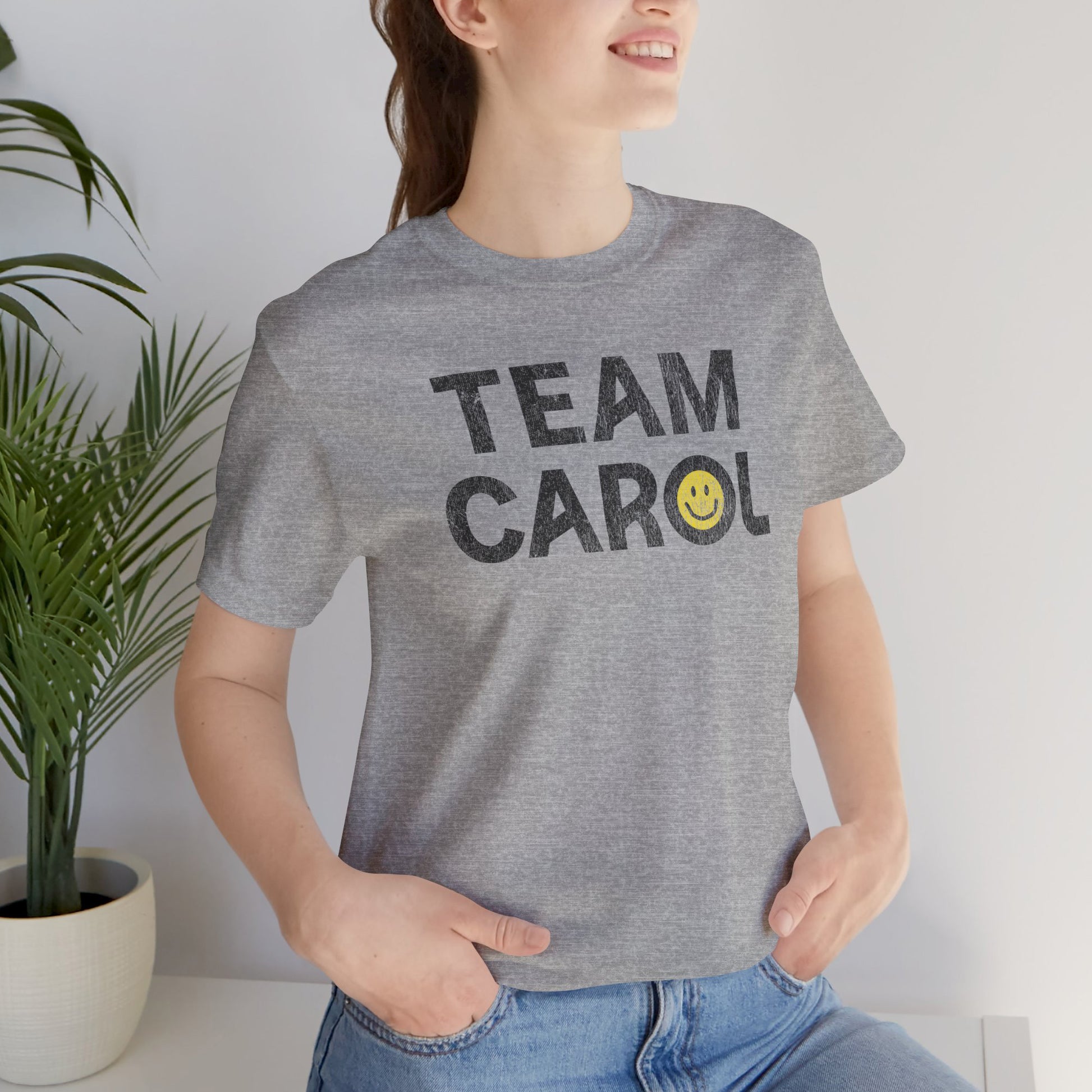 Person wearing heather gray TEAM CAROL t-shirt inspired by Pluribus with vintage distressed smiley face graphic, seated near a plant